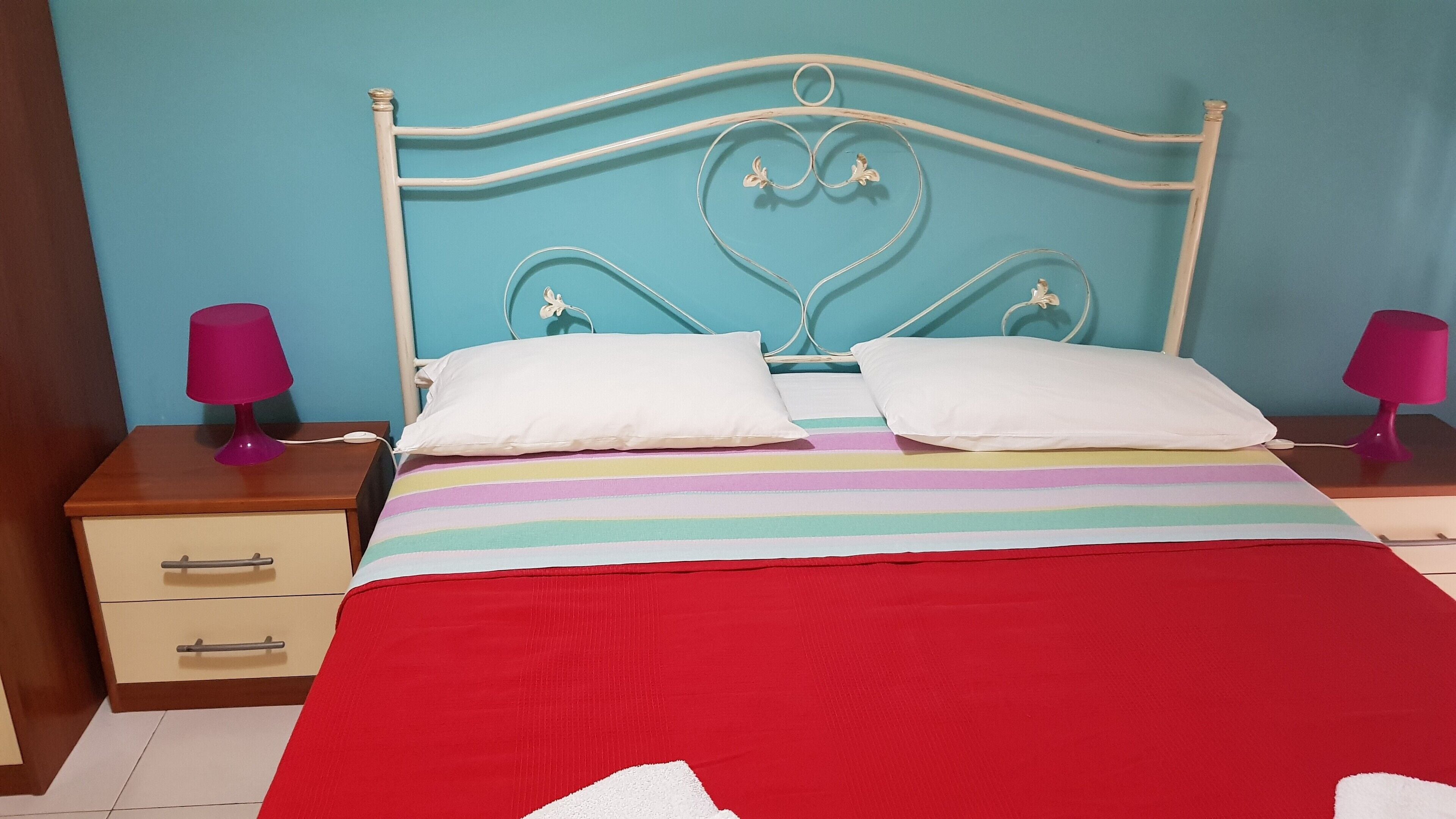 A bed with a striped bedspread and a red blanket, two white pillows, a white headboard with heart-shaped designs, and two bedside tables with lamps.