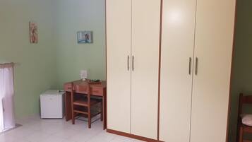 A room with a large wardrobe, a desk with a chair, a small refrigerator, and a rug on the floor.