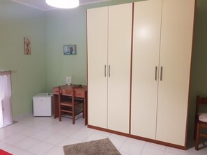 A room with a large wardrobe, a desk with a chair, a small refrigerator, and a rug on the floor. - The Artists (Acireale)