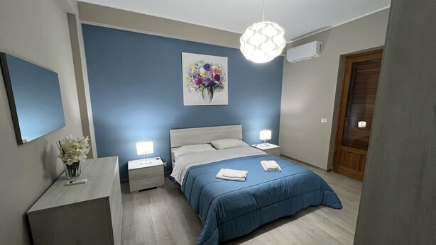 A modern bedroom with a blue accent wall, a wooden headboard, a bed with blue bedding, two bedside tables with lamps, a small desk with a vase of flowers, and a painting above the bed. - The Artists (Acireale)