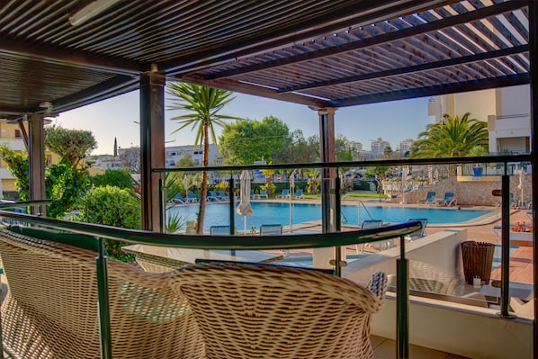 A balcony with wicker chairs overlooking a swimming pool and a cityscape.