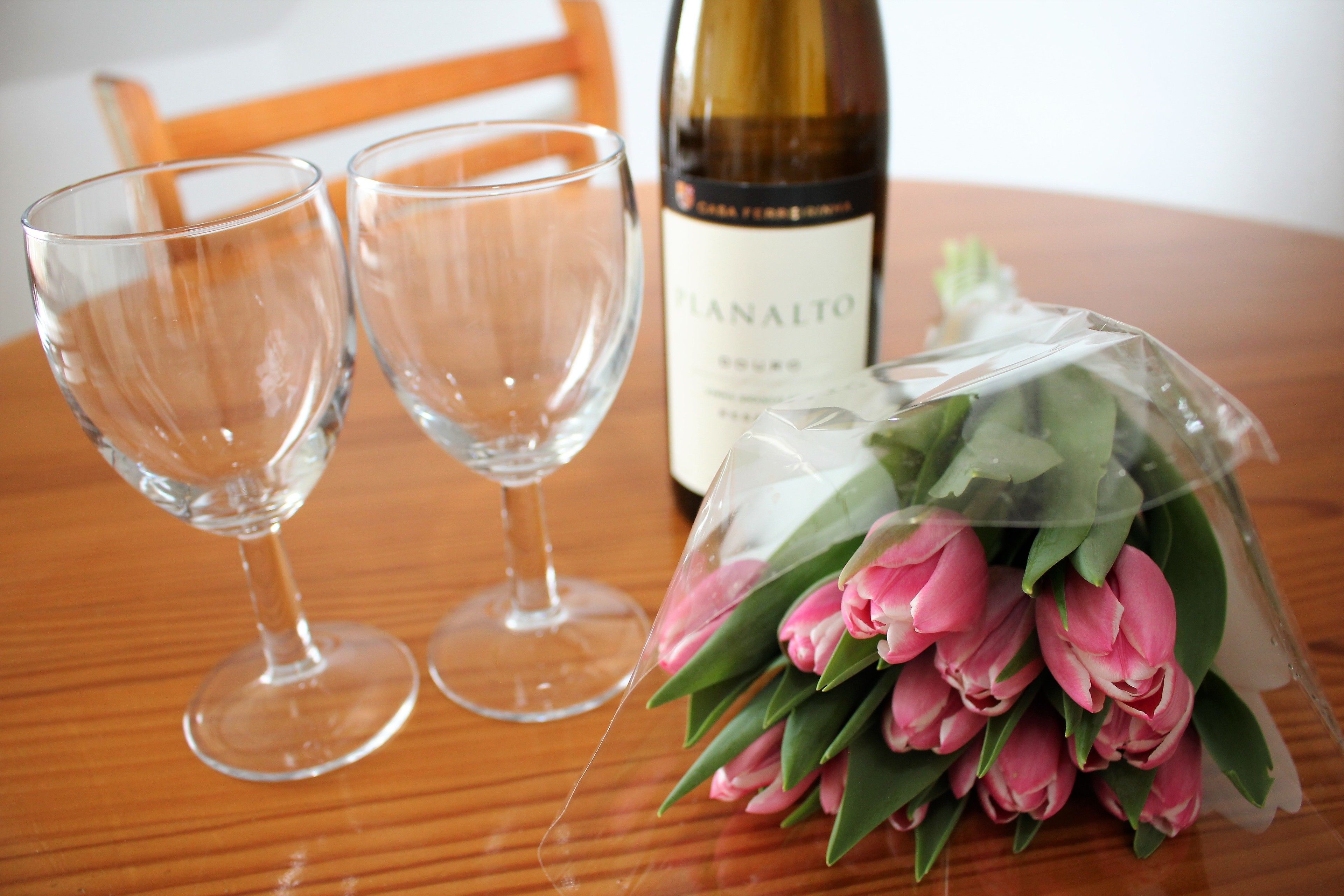 Deux verres à vin vides, une bouteille de vin et un bouquet de tulipes roses sur une table en bois.