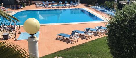 A swimming pool with lounge chairs and a sun lamp, surrounded by greenery and buildings.