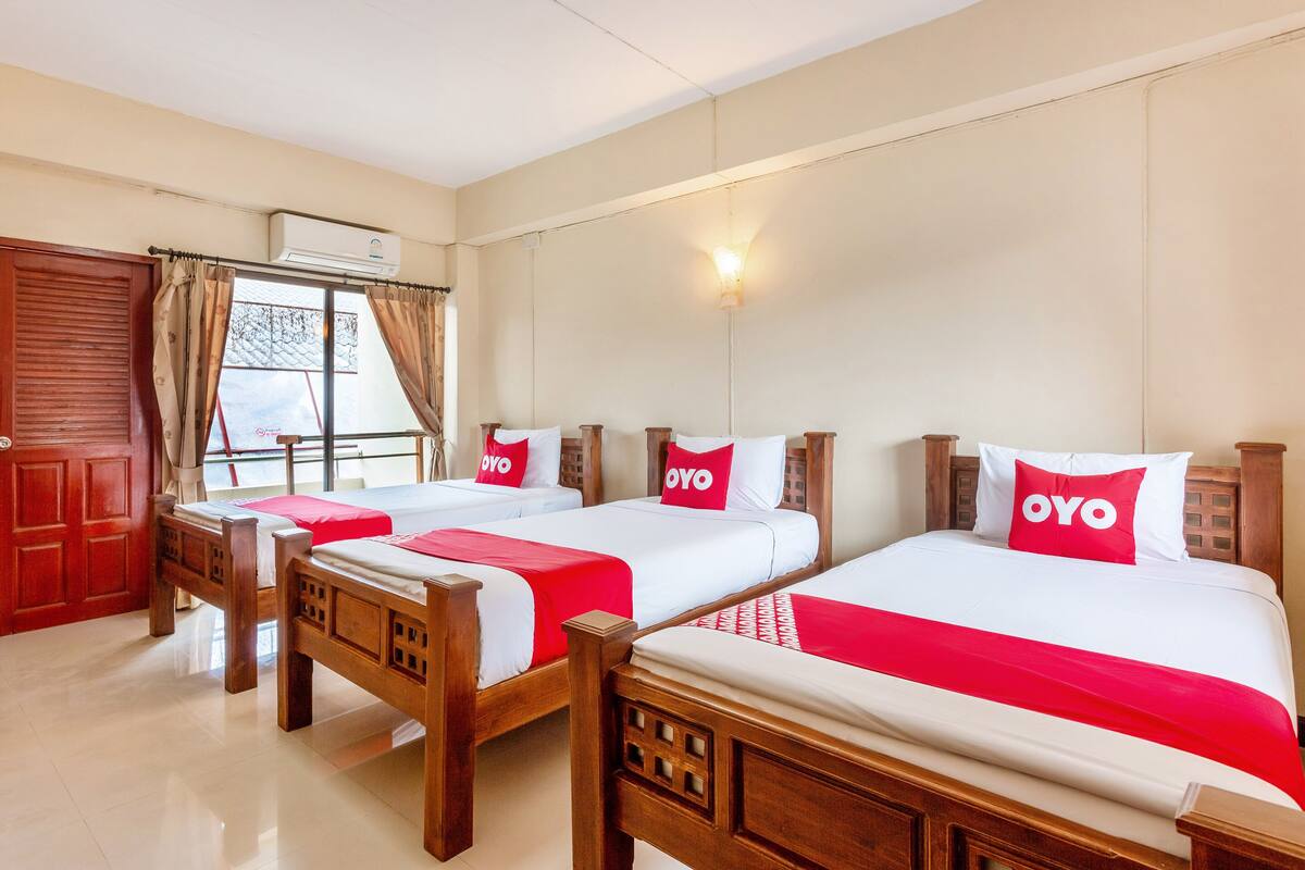 A hotel room with three wooden beds, each with white bedding and red accents, a red door, and a window with curtains.