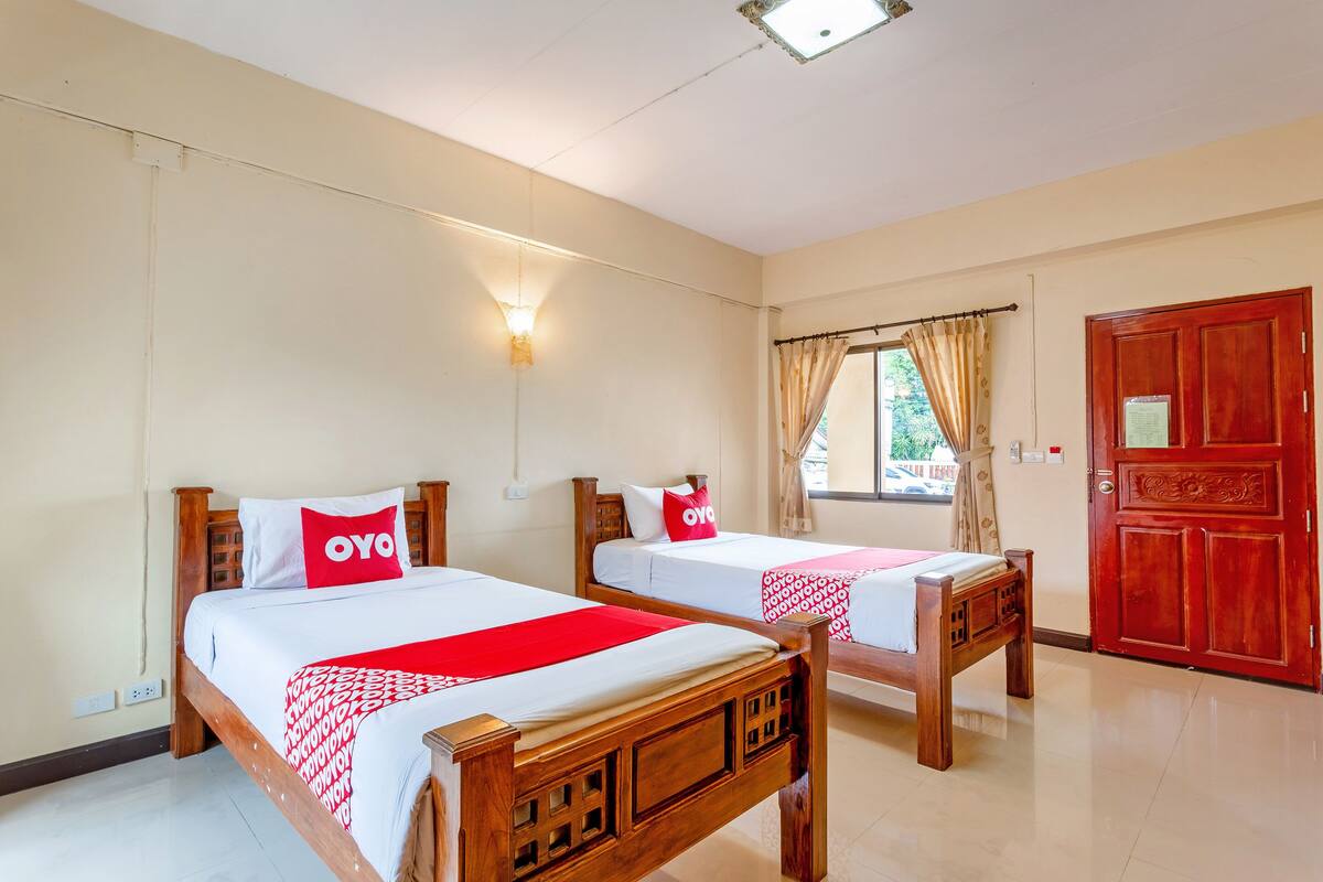 A hotel room with two wooden beds, each with a red and white bedspread, a red pillow with the logo "OYO", a wall-mounted light, and a red door.
