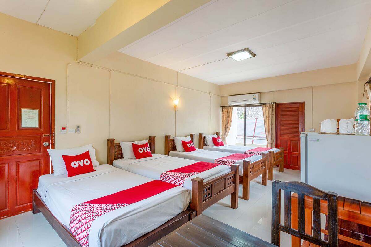 A hotel room with three beds, each with a red OYO pillow, wooden headboards, and a white bedspread. There is a wooden desk with a chair, a white refrigerator, and a window with curtains.