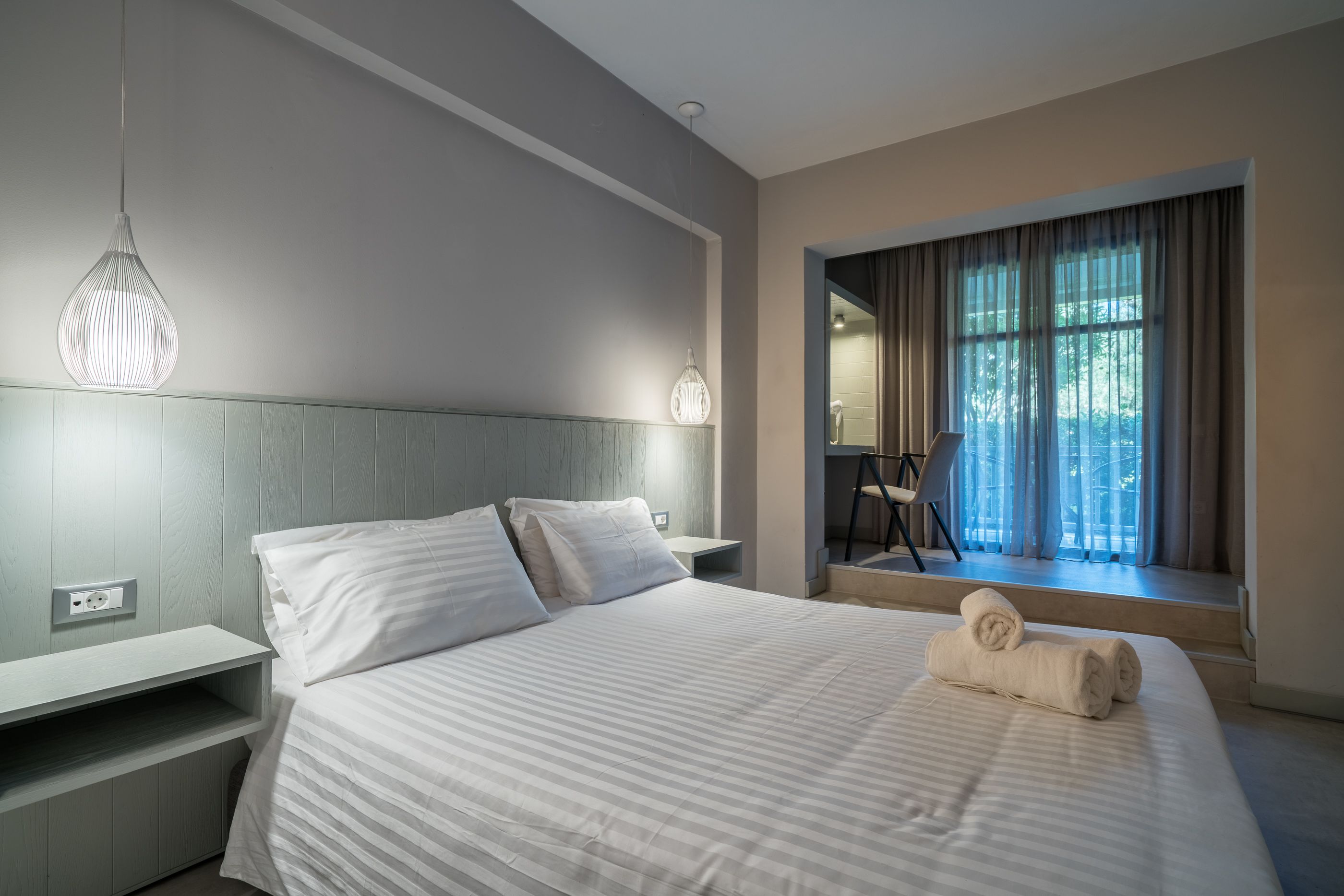 Una habitación de hotel moderna con una cama grande, una mesita de noche, una lámpara colgante y un ventanal con cortinas.