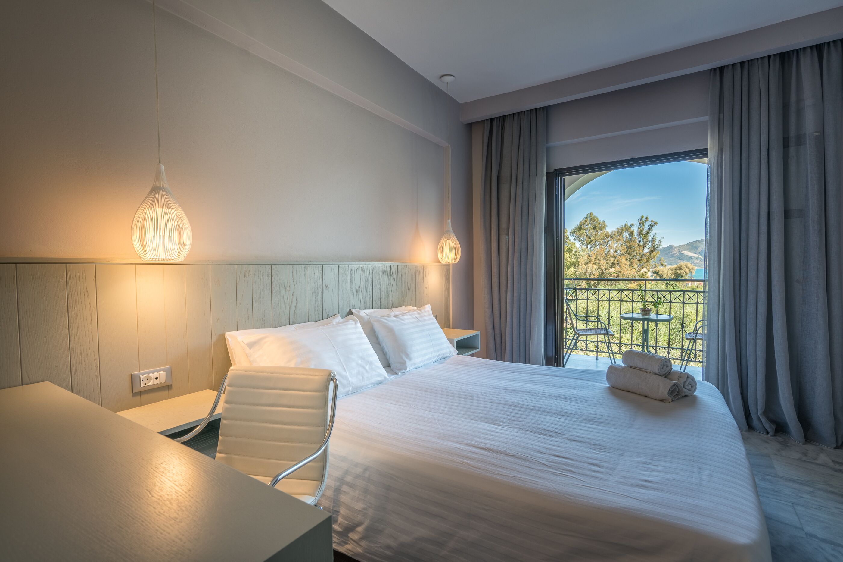 Habitación de hotel con cama, escritorio, silla, lámpara colgante y balcón con vista al exterior.