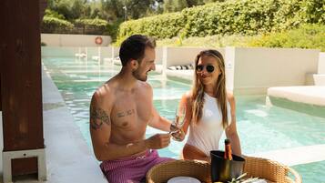 A poolside setting with a wicker basket containing food and drinks, a man and a woman, and a lifebuoy in the background.