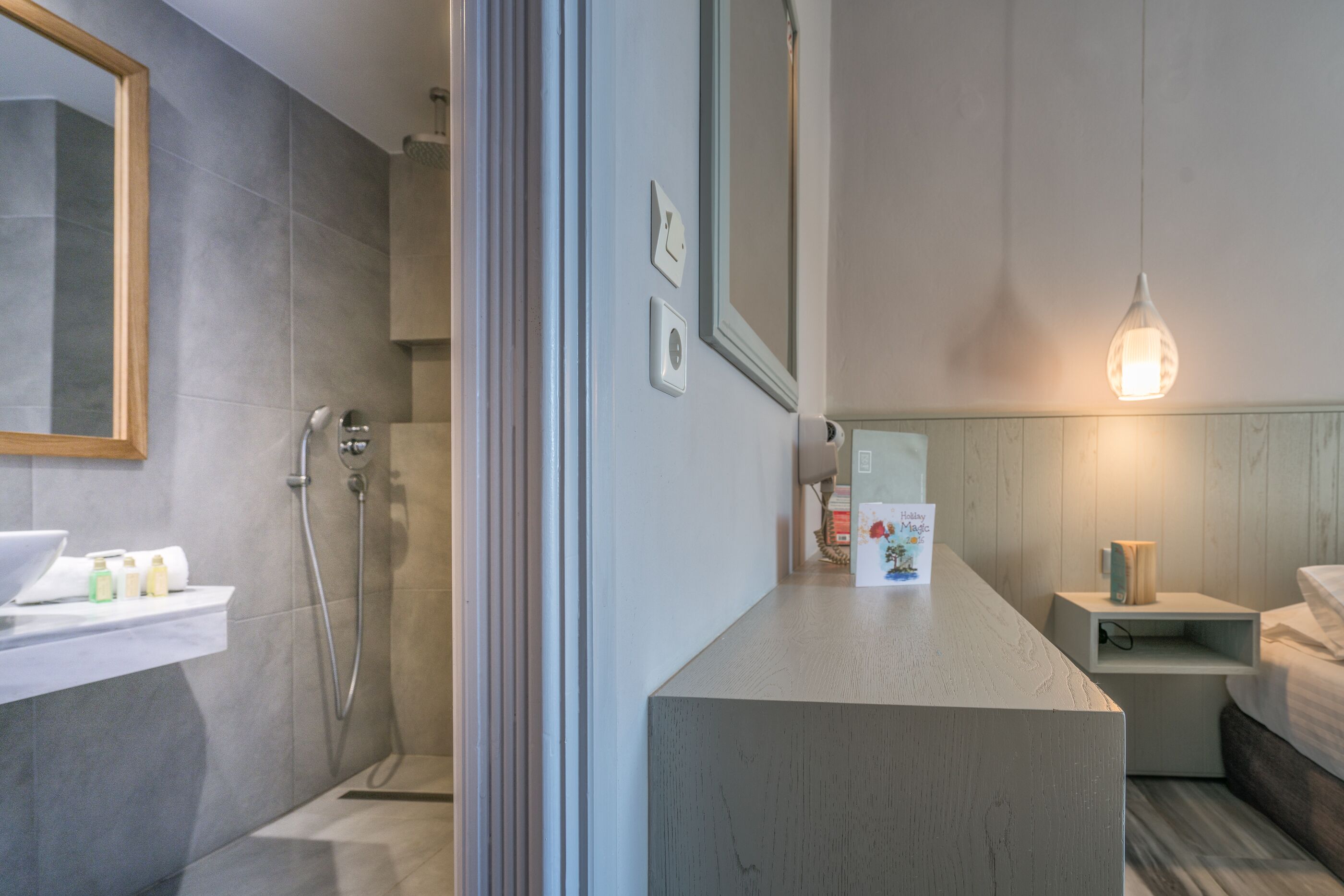 A modern bathroom with a shower, a sink with toiletries, a mirror, and a bedside table with a lamp.
