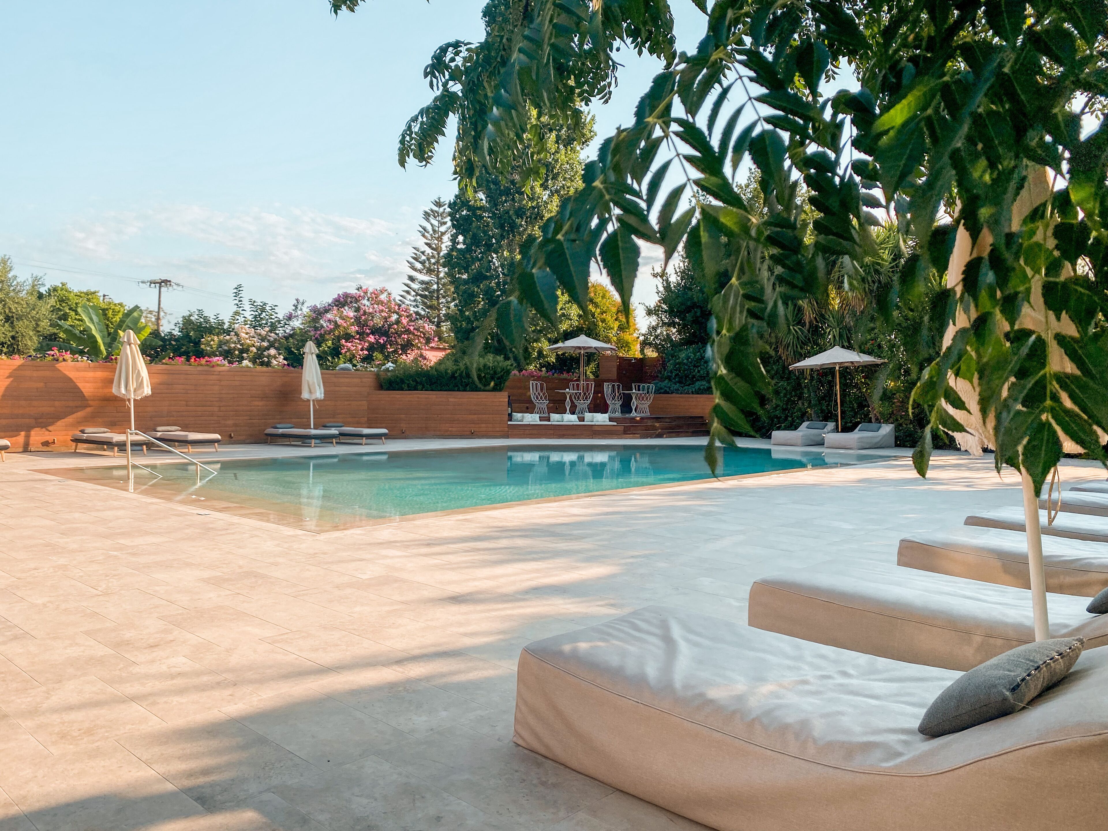 Zona de piscina con sillas de descanso y sombrillas, rodeada de vegetación y un muro de ladrillo.