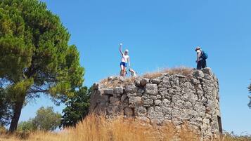 Ruinas de piedra con dos personas de pie en la cima, rodeado de pasto seco y árboles.