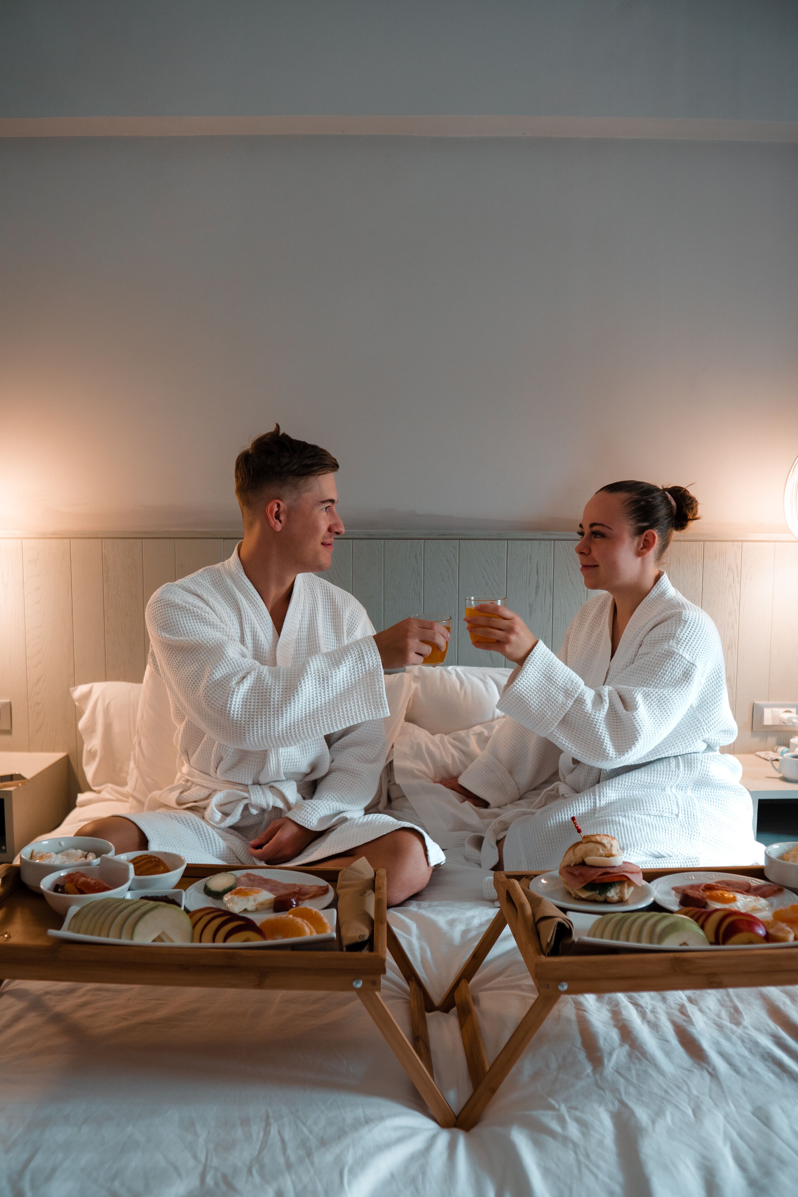A couple in white bathrobes sitting on a bed with breakfast trays.