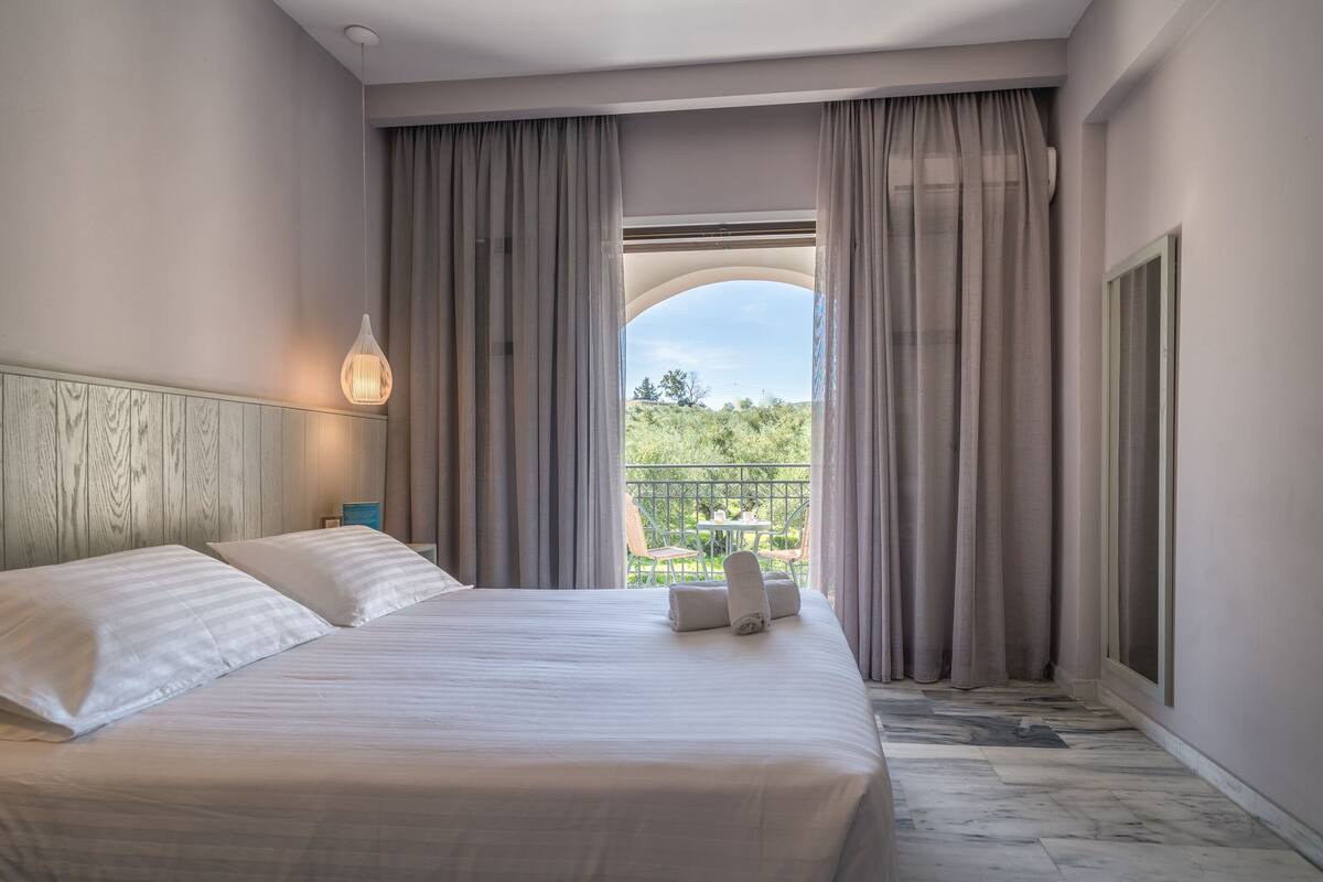 Una camera d'albergo con un letto grande, un comodino, una lampada a sospensione e vista su un balcone con piante.
