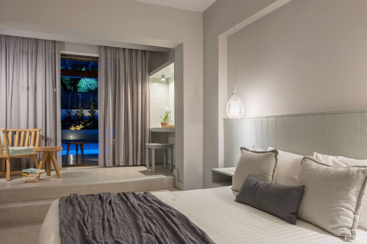 Una moderna camera d'albergo con un letto grande, un'area salotto con una sedia e un tavolo, e vista sull'esterno tramite una porta vetrata.