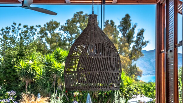 An outdoor seating area with tables and chairs, a wicker hanging light fixture, and a view of greenery and a mountain in the background. - Castelli Hotel Adults Only - All Inclusive (Zakynthos)