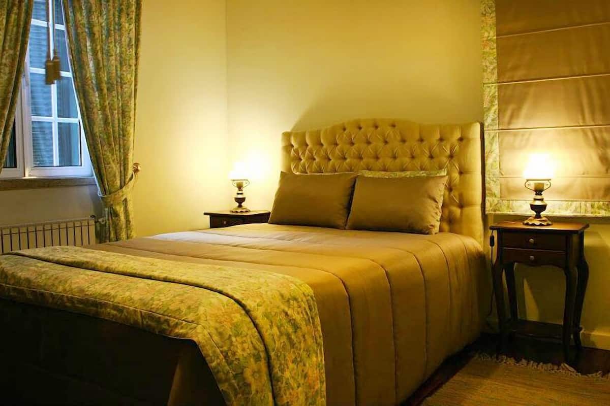 A bedroom with a tufted headboard, two bedside tables with lamps, a window with curtains, and a radiator.