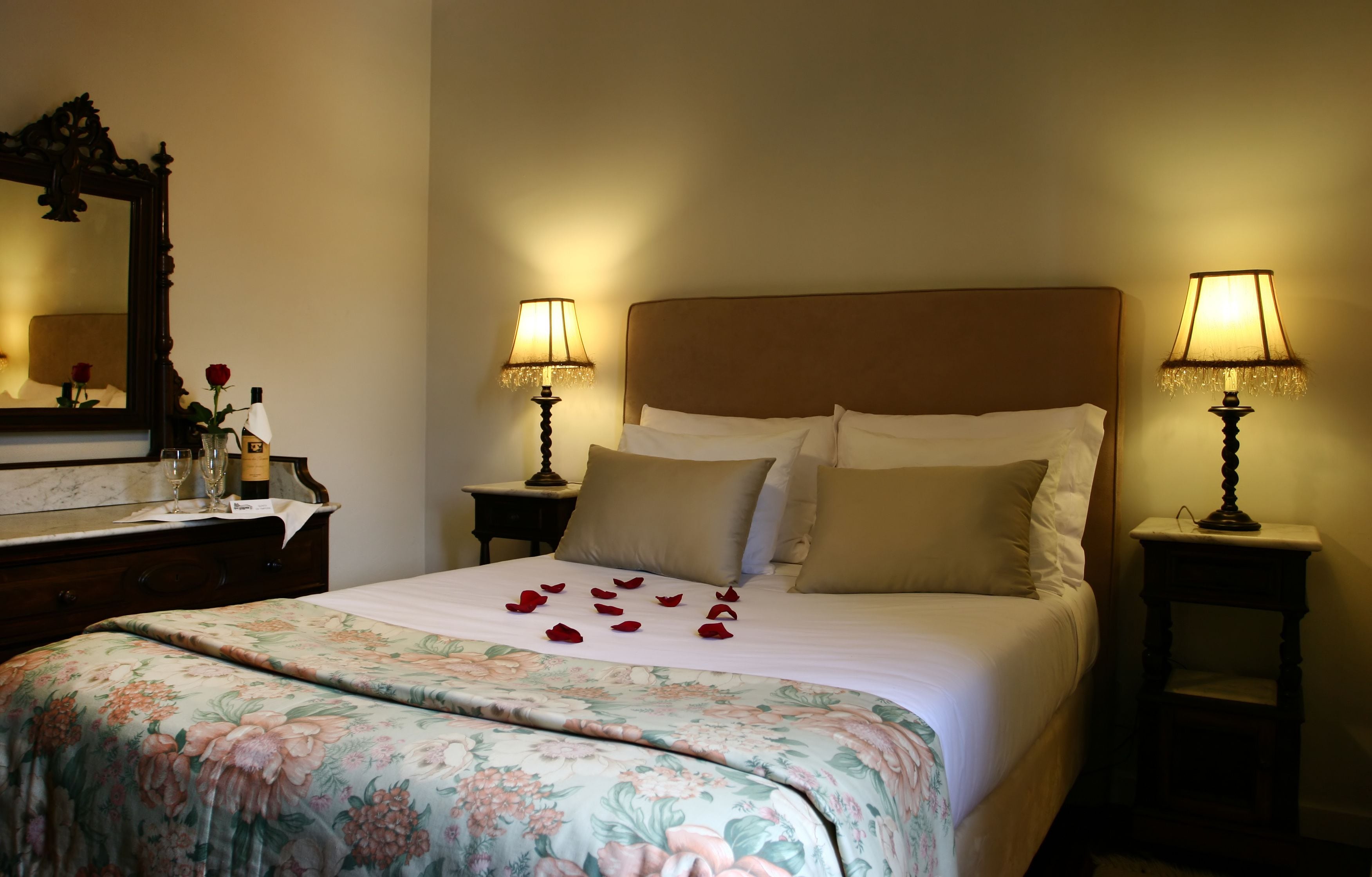 A hotel room with a bed, two bedside lamps, a nightstand, a mirror, and a floral bedspread.