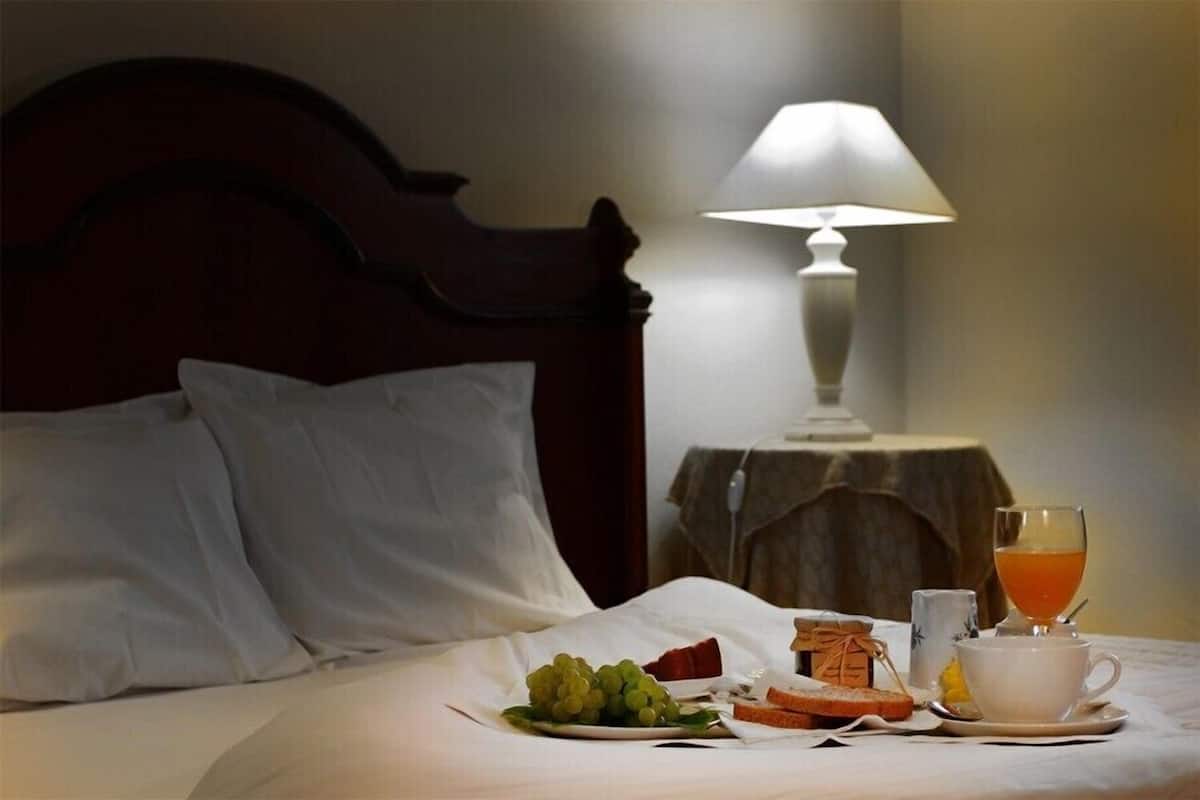 A neatly made bed with white linens, a bedside table with a lamp, and a breakfast tray with toast, grapes, and a cup of tea.
