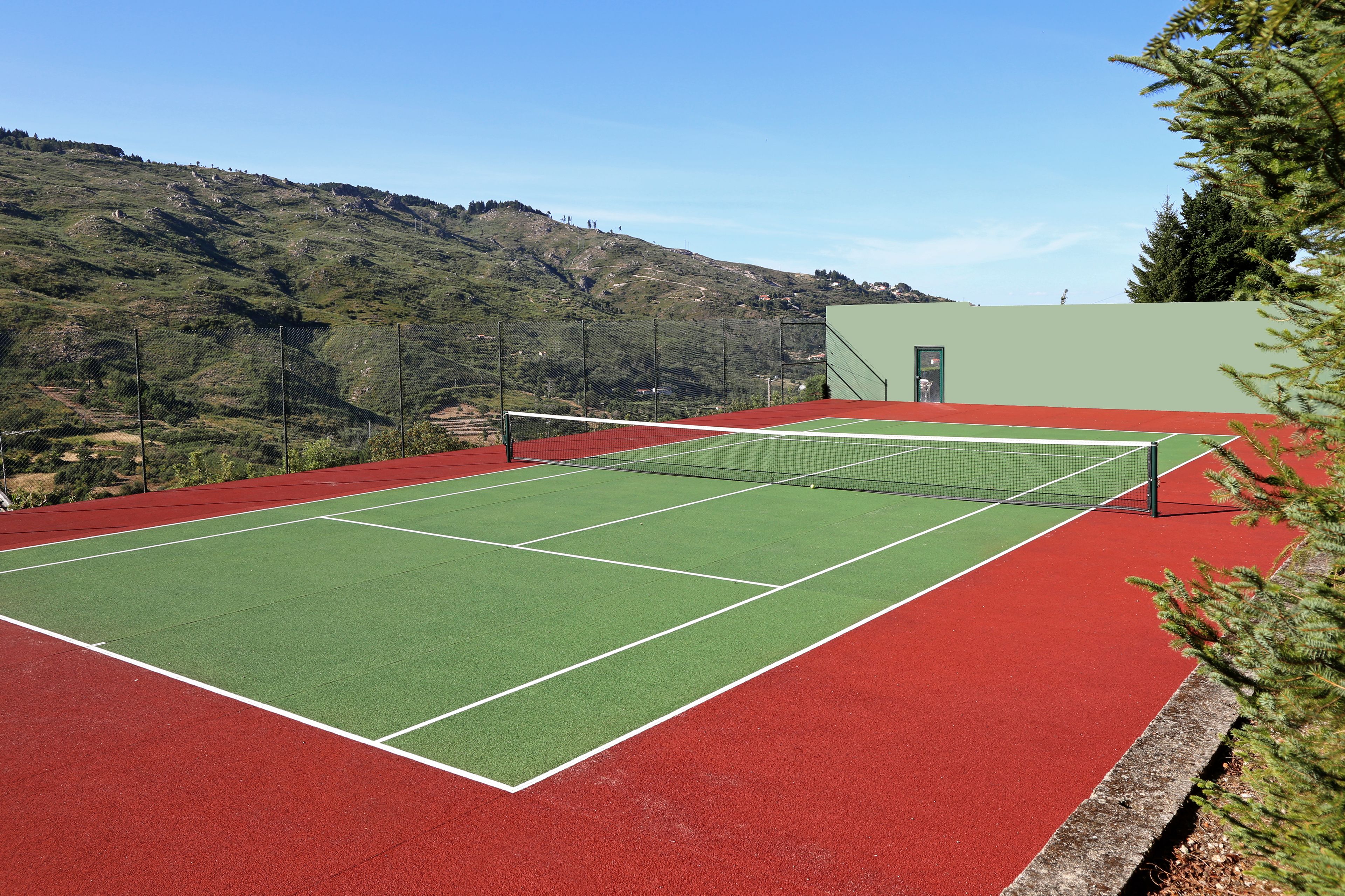 Un court de tennis avec un filet, entouré d’une surface rouge et verte, et un paysage montagneux en arrière-plan.