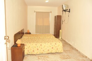 A small, narrow room with a bed, a nightstand, a lamp, and a wall-mounted heater.