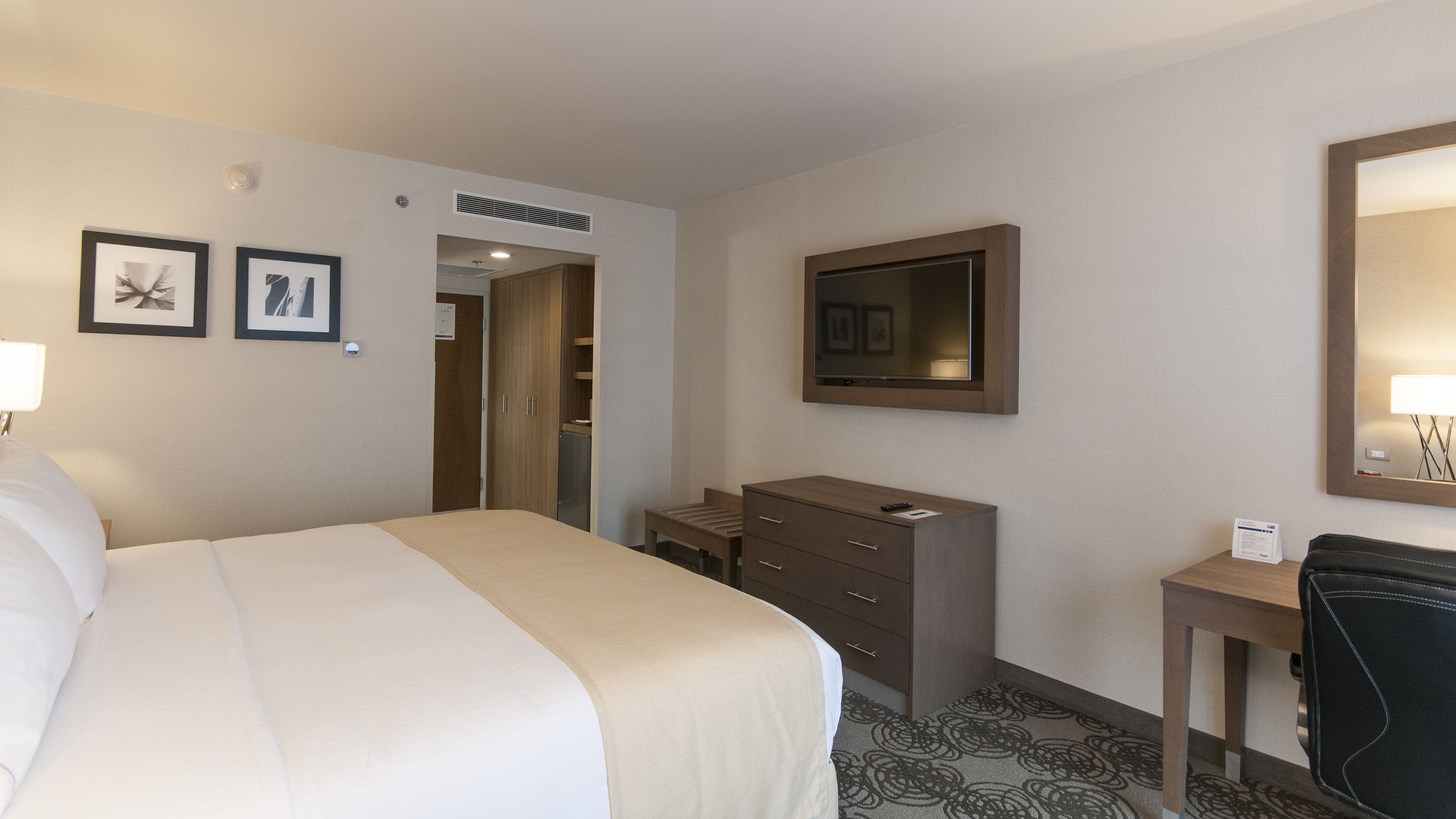 A hotel room with a bed, a dresser, a desk, a mirror, and framed pictures on the wall.