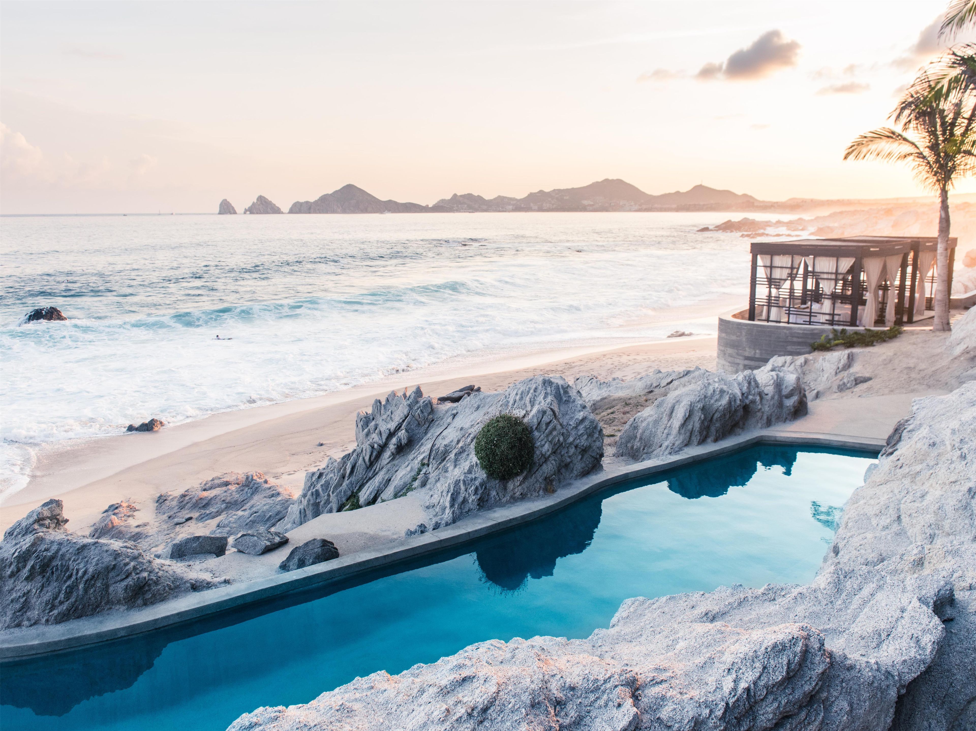 Una piscina junto a una playa, con vista al océano y montañas distantes.