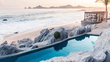 Una piscina junto a una playa, con vista al océano y montañas distantes.