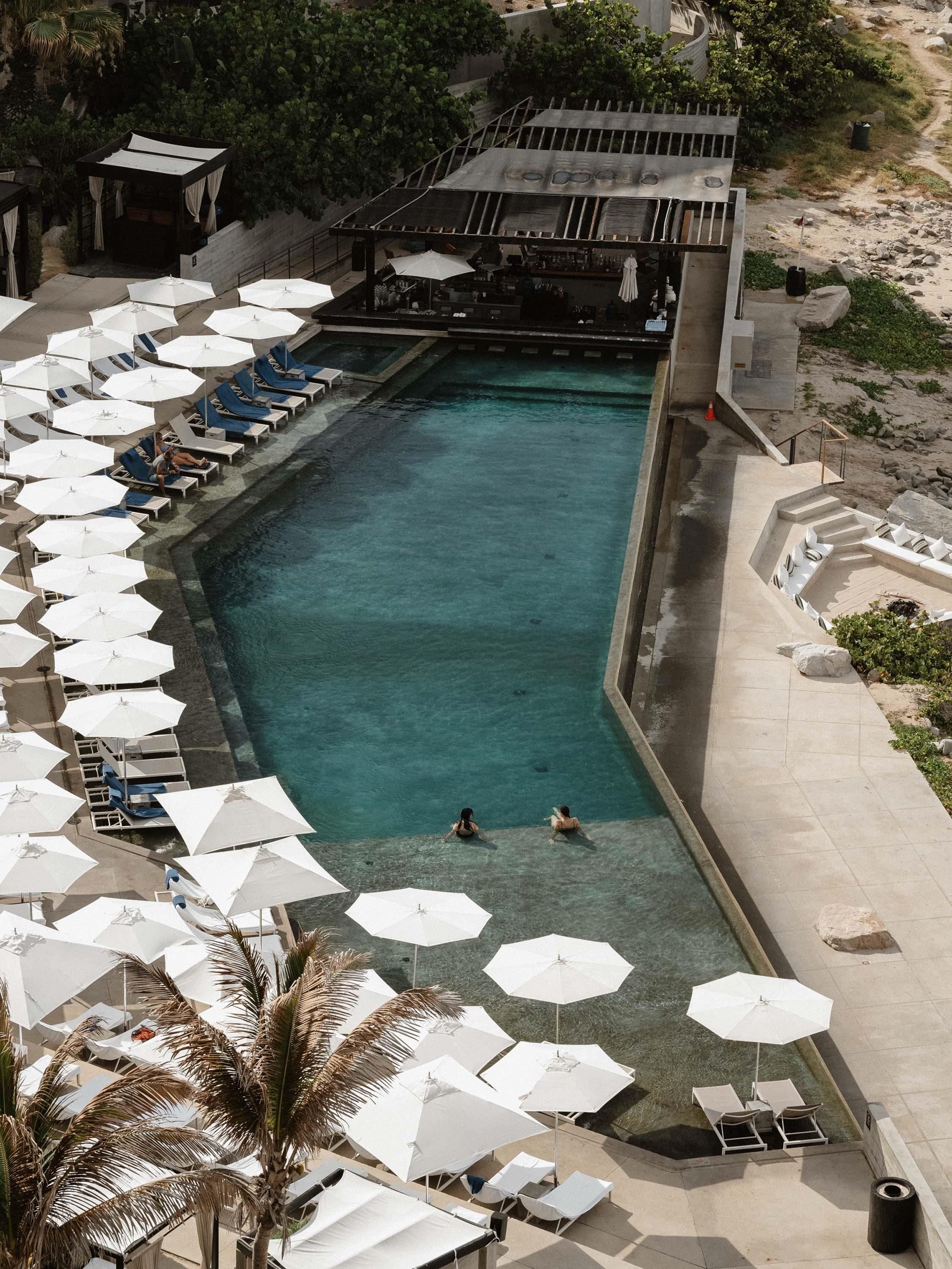 A swimming pool with lounge chairs and umbrellas, surrounded by a concrete deck and a covered area with a bar.
