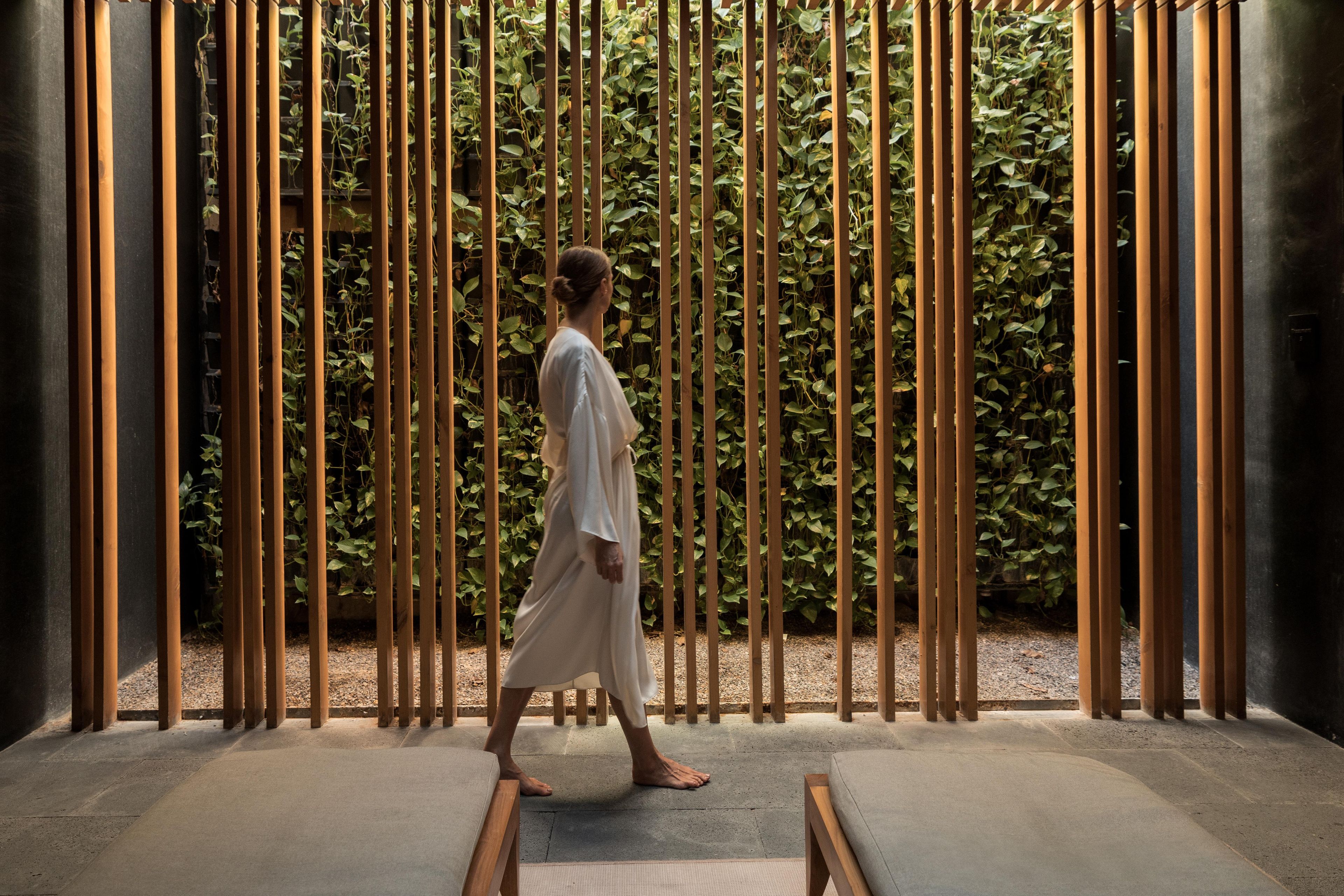 A person walking through a space with vertical wooden slats and a green leafy background.