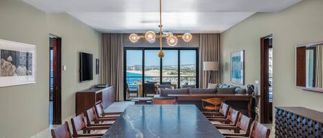 A spacious dining area with a long table and chairs, a modern chandelier, and a view of the sea through large windows.