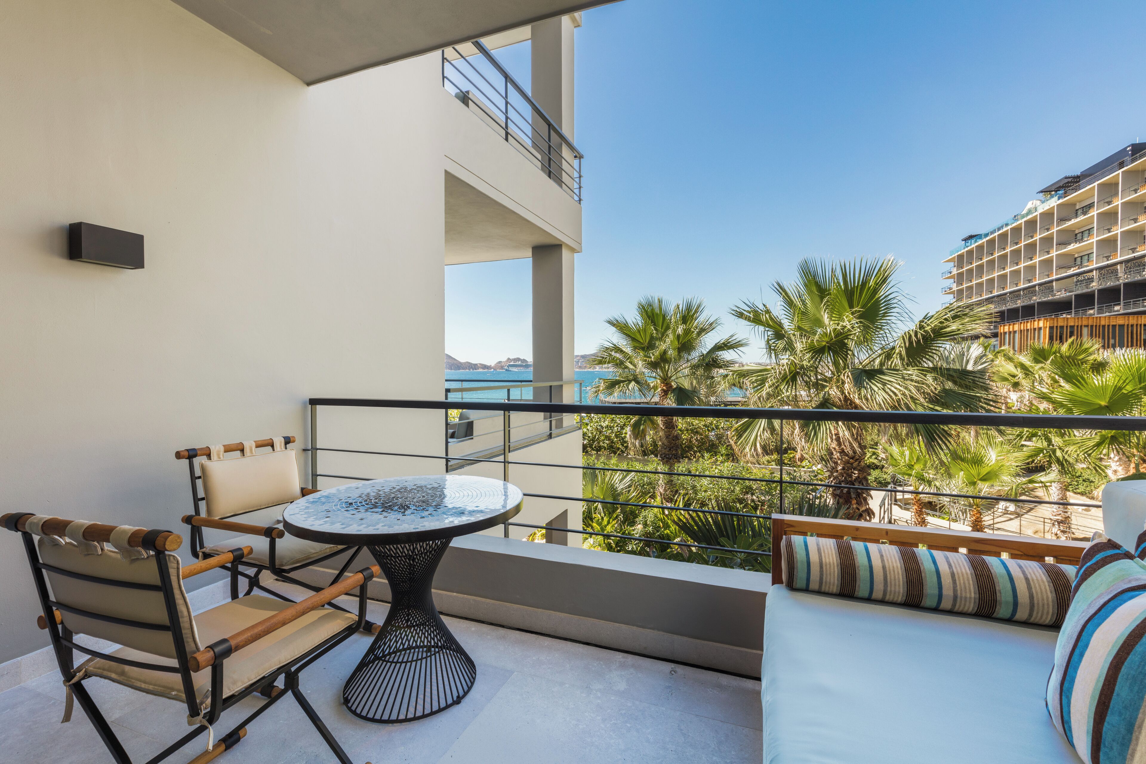 A balcony with a round table and chairs, a sofa with striped cushions, and a view of palm trees and a building across the water.