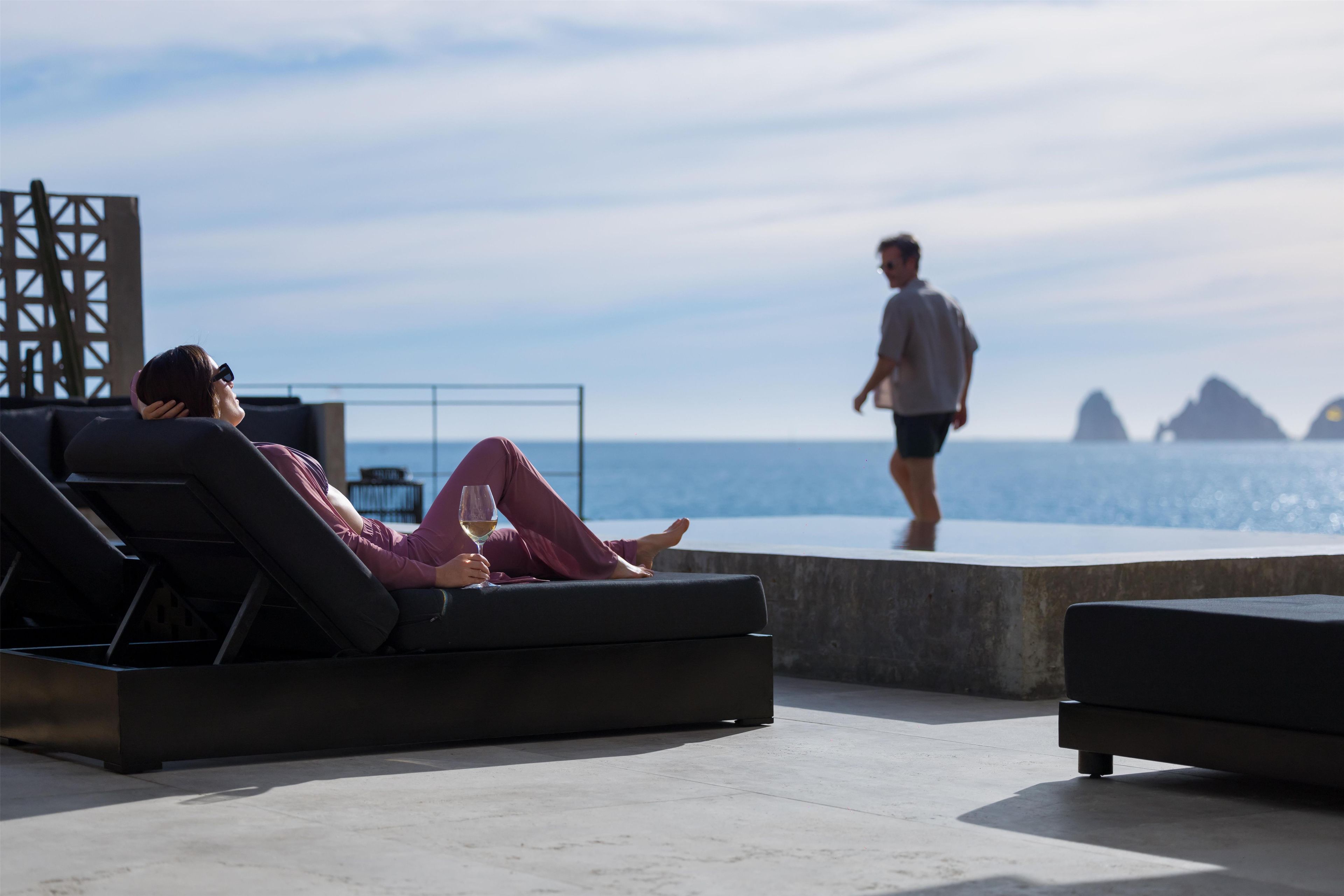Une personne allongée sur une chaise longue, un verre à la main, et qui contemple un plan d’eau ainsi que des reliefs lointains.