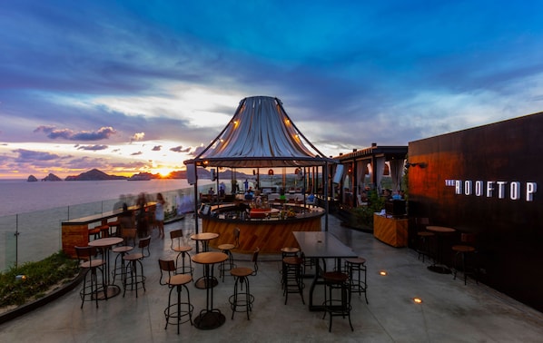 An outdoor bar area with a view of the sunset over the water, tables, chairs, and a sign reading "THE ROOFTOP". - The Cape, A Thompson Hotel, by Hyatt (Cabo San Lucas)