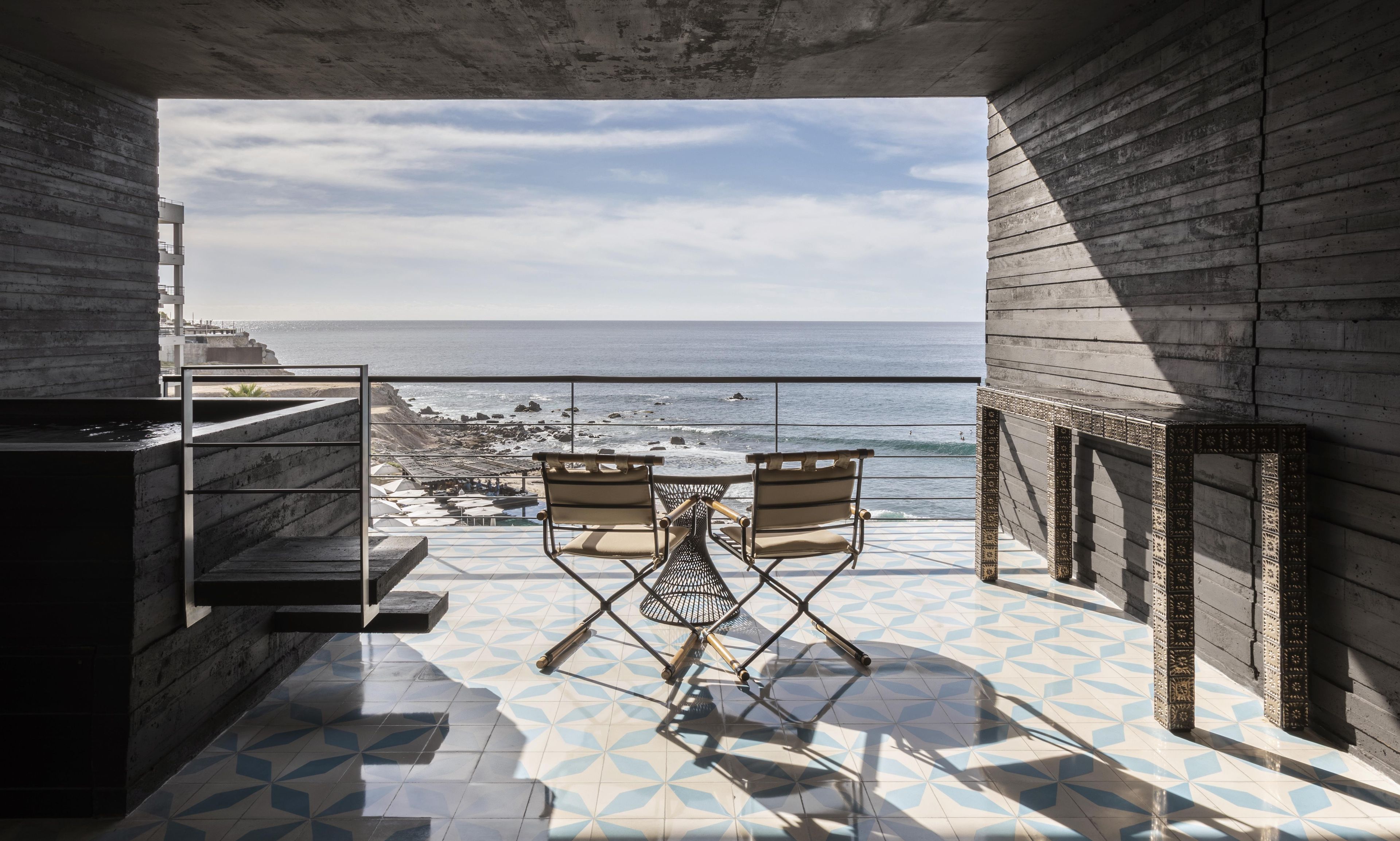 A balcony with two chairs facing a beach, a tiled floor, and a view of the ocean.