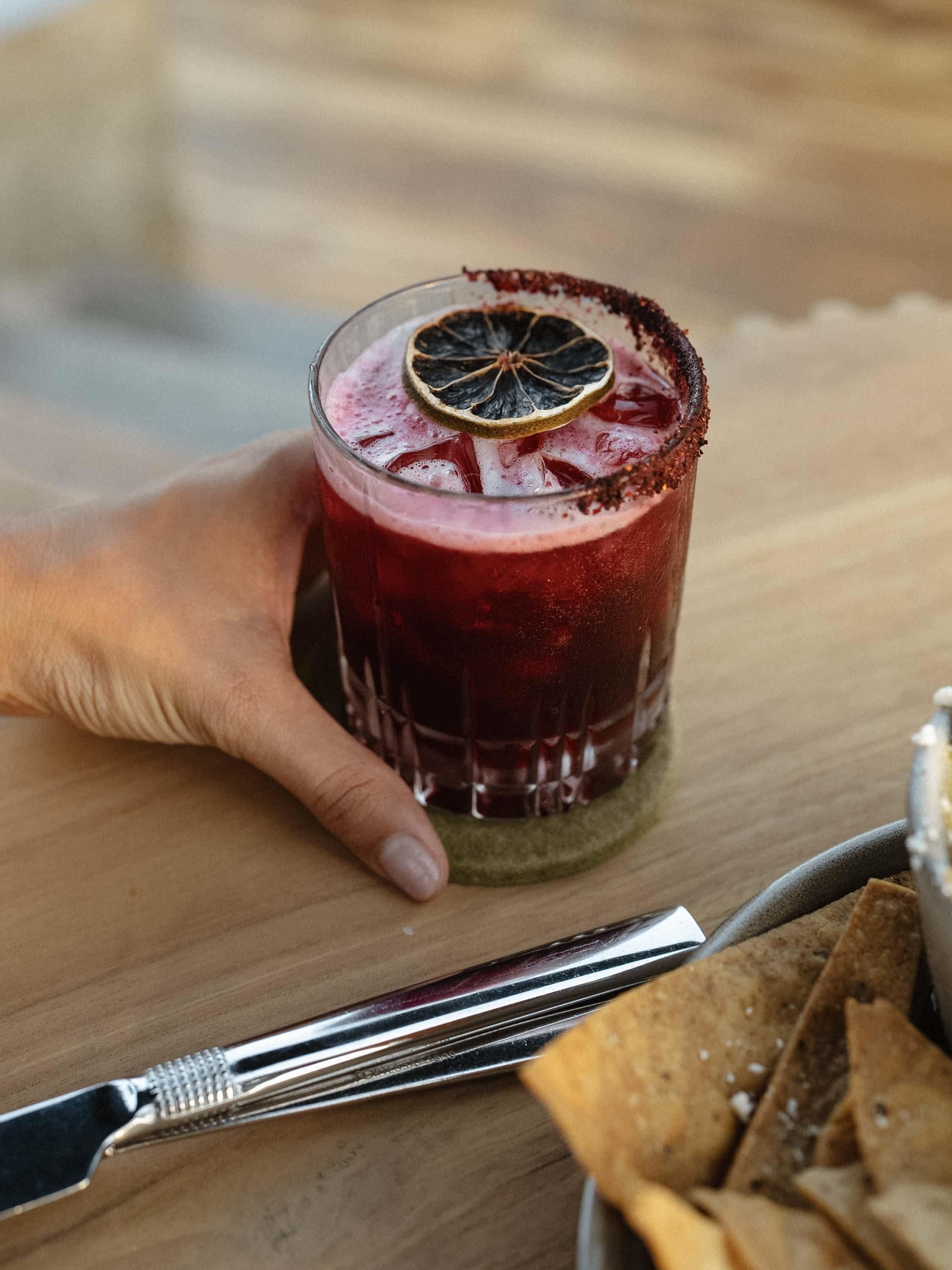 A hand holding a glass with a dark red beverage, garnished with a slice of dried fruit and a rim coated with a dark substance, possibly chocolate or spice, on a wooden surface.