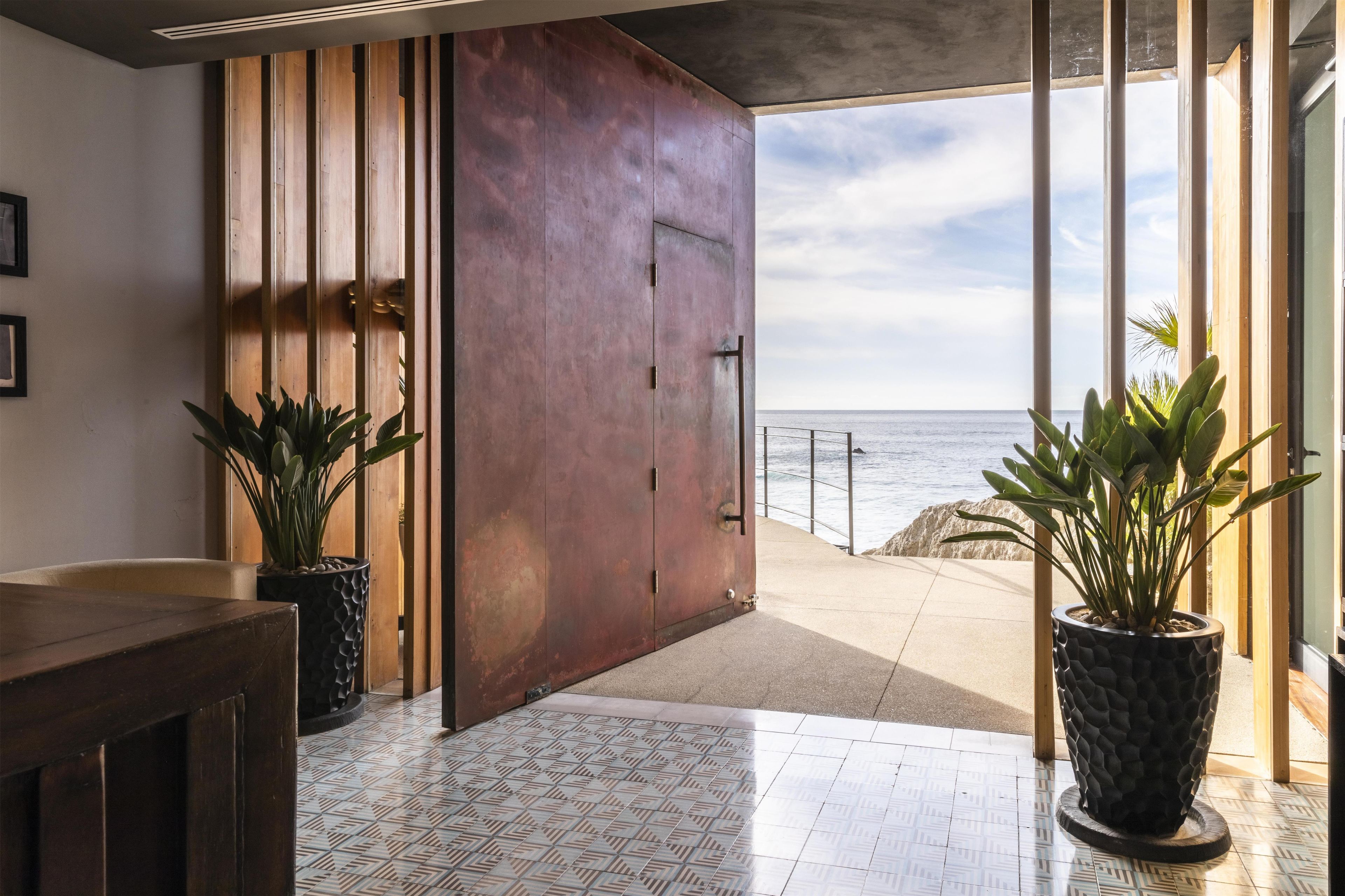 A modern interior with a large wooden door leading to a balcony with a view of the sea. There are potted plants and a tiled floor.