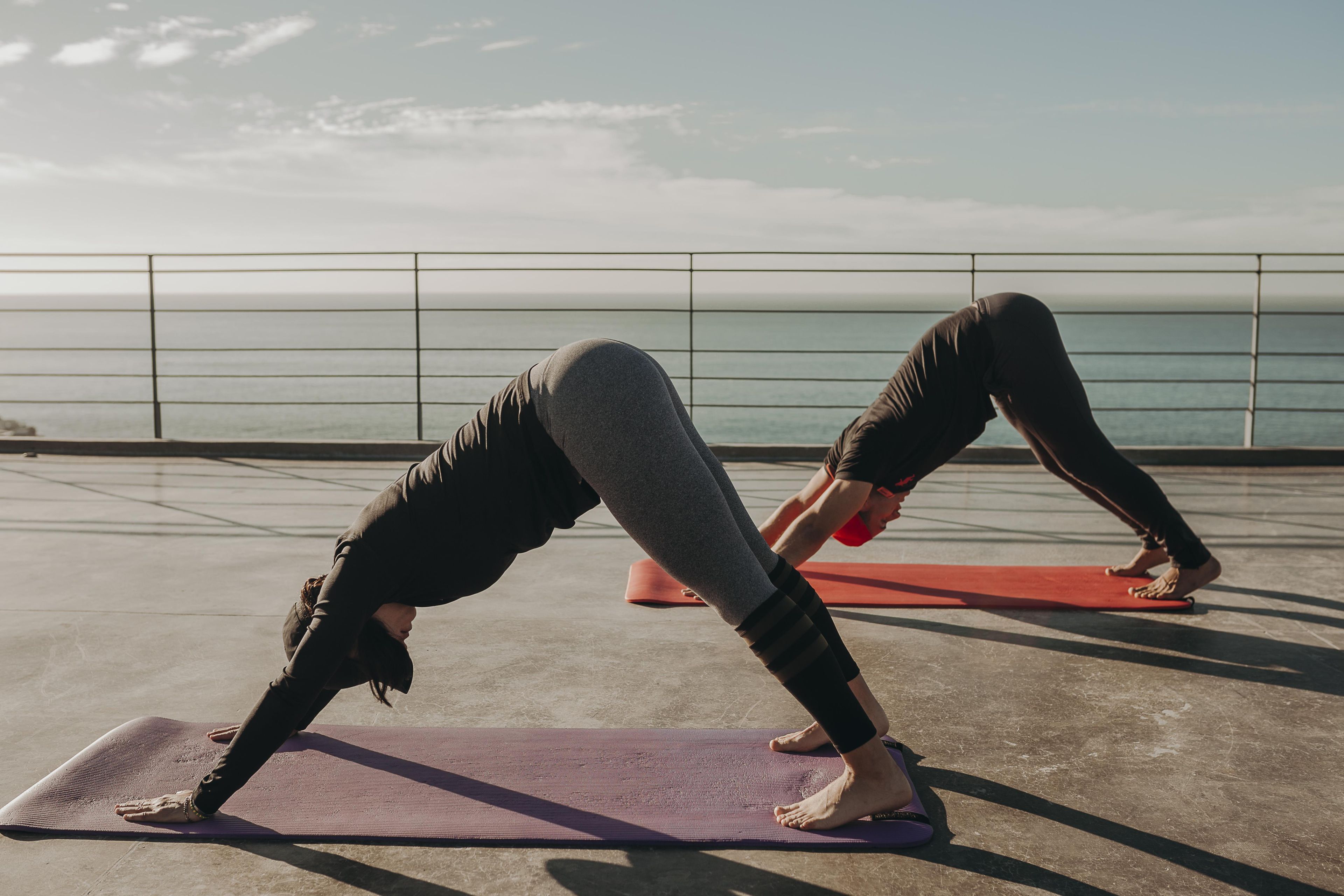 Twee mensen doen yoga op matten op een betonnen vloer, met een reling en een waterlichaam op de achtergrond.
