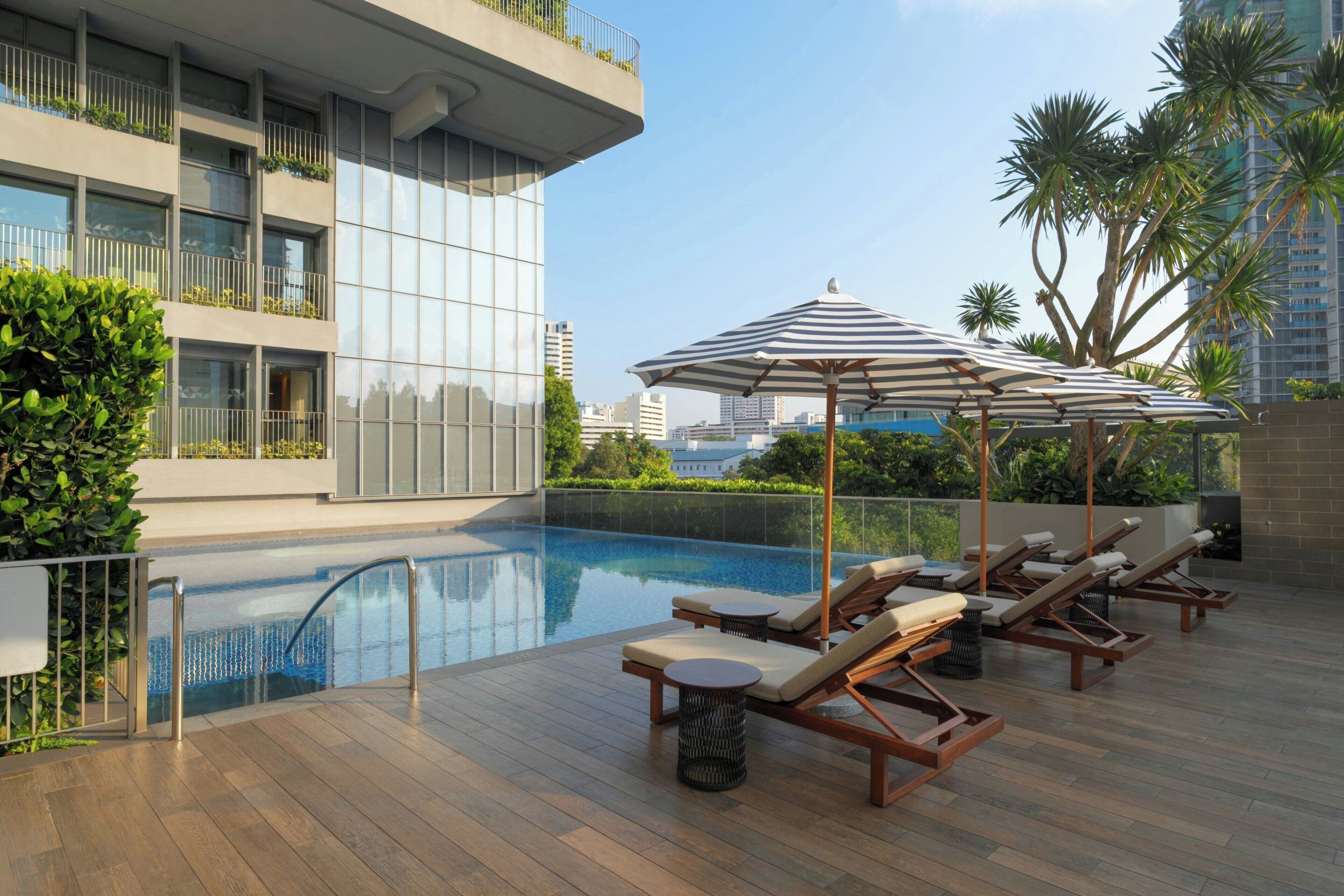 A rooftop pool area with lounge chairs and umbrellas, surrounded by greenery and modern buildings.