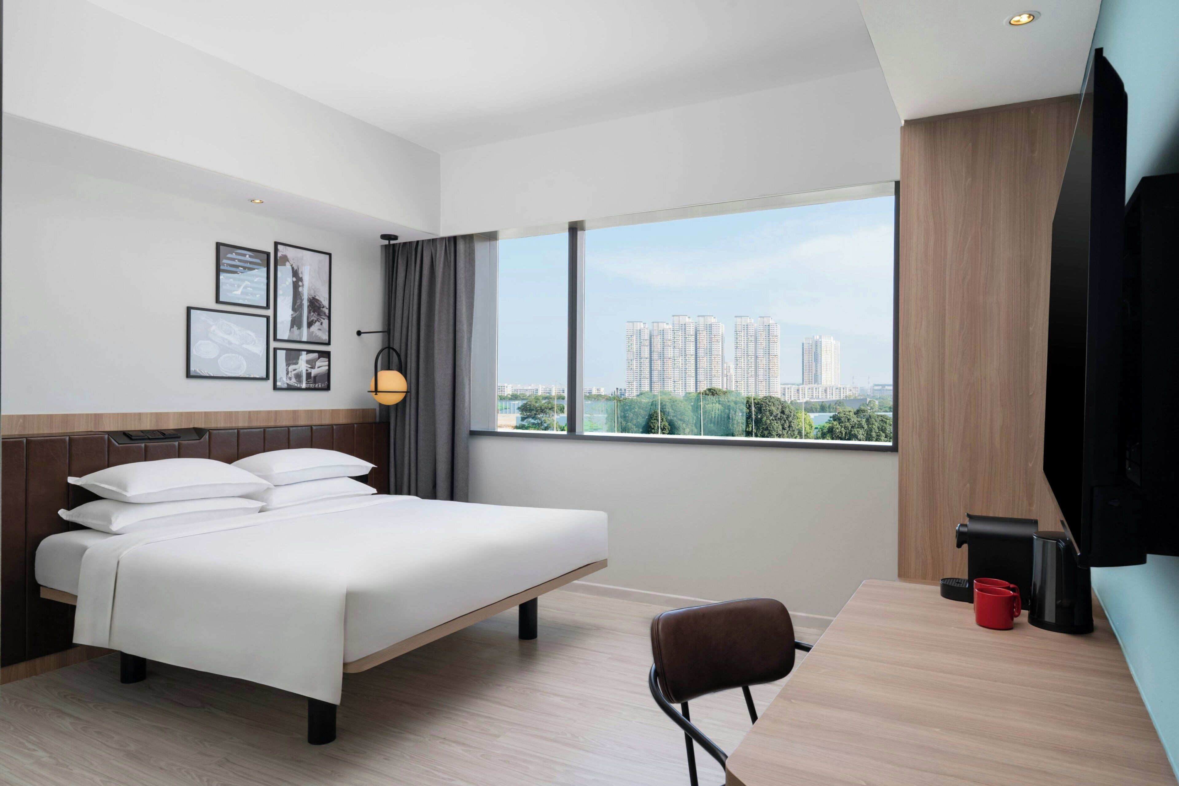 Une chambre d’hôtel avec un lit, un bureau, une chaise, une fenêtre donnant sur la ville et des tableaux encadrés accrochés au mur.
