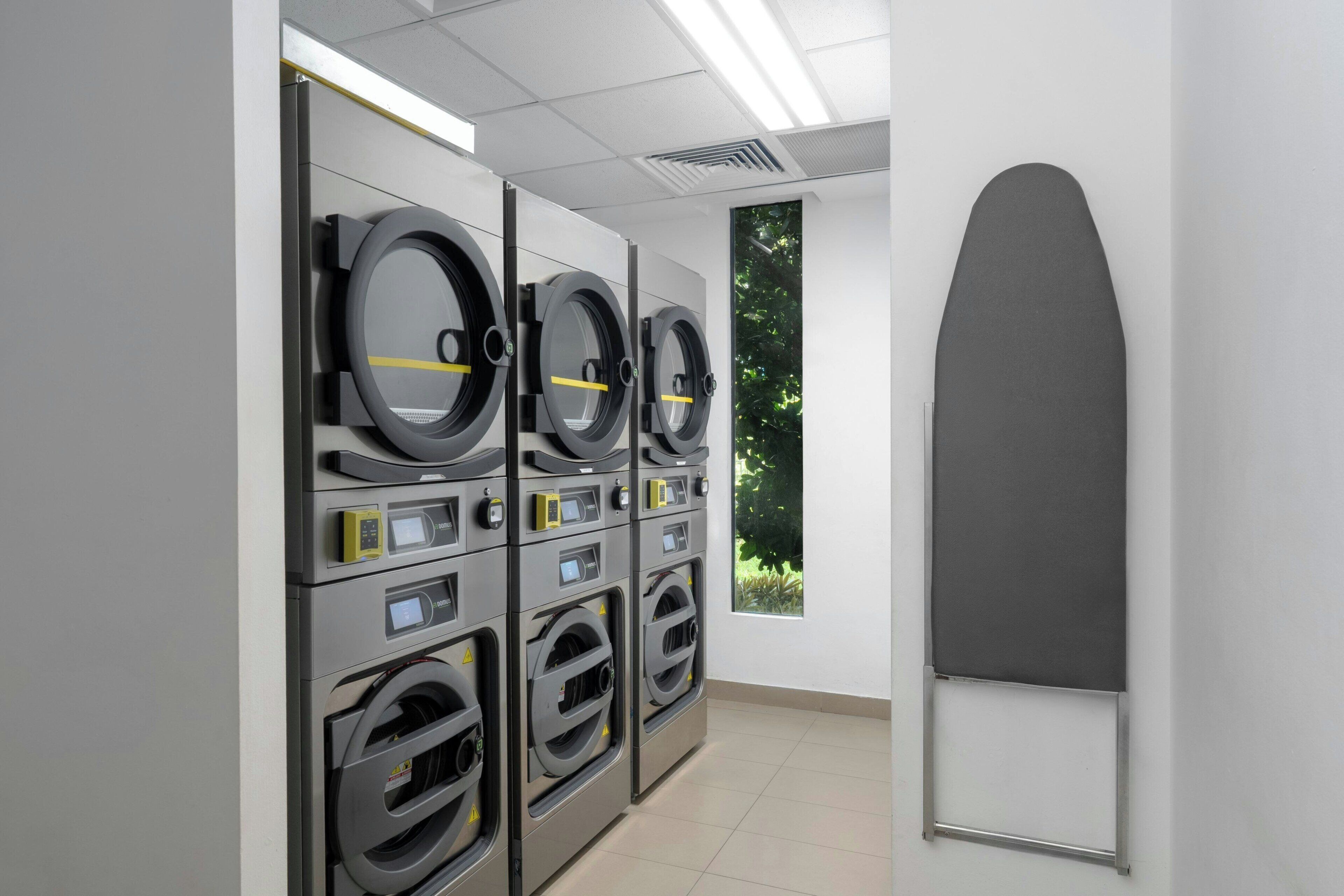 A row of industrial-style laundry machines in a clean, well-lit room with a view of greenery through a window.