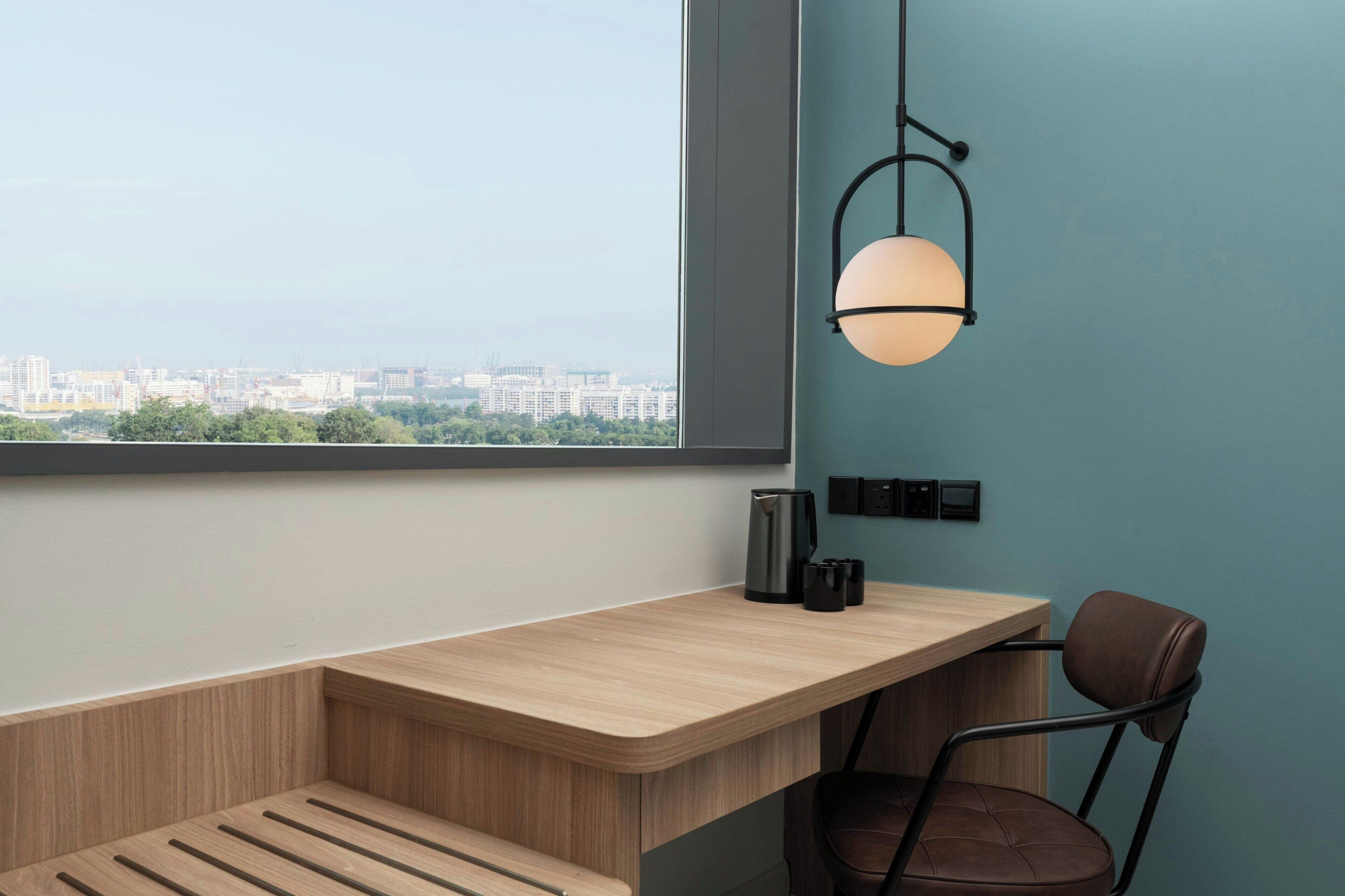 A modern room with a wooden desk, a chair, a hanging light fixture, and a view of a cityscape through a large window.