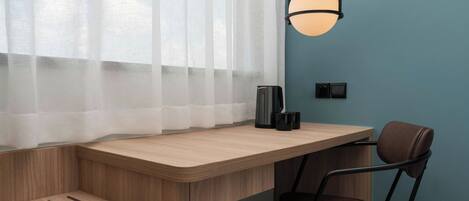 Una habitación moderna con un escritorio de madera, una silla, un ventanal con cortinas translúcidas y una lámpara fijada a la pared.