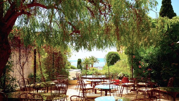 An outdoor patio with metal chairs and tables, surrounded by lush greenery and a view of the sea. - Hotel Royal Bon Repos (Sainte-Maxime)