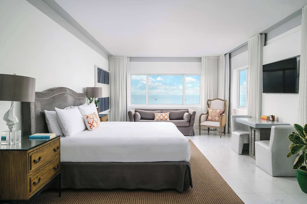 A modern hotel room with a large bed, a desk, a chair, and a view of the sea.