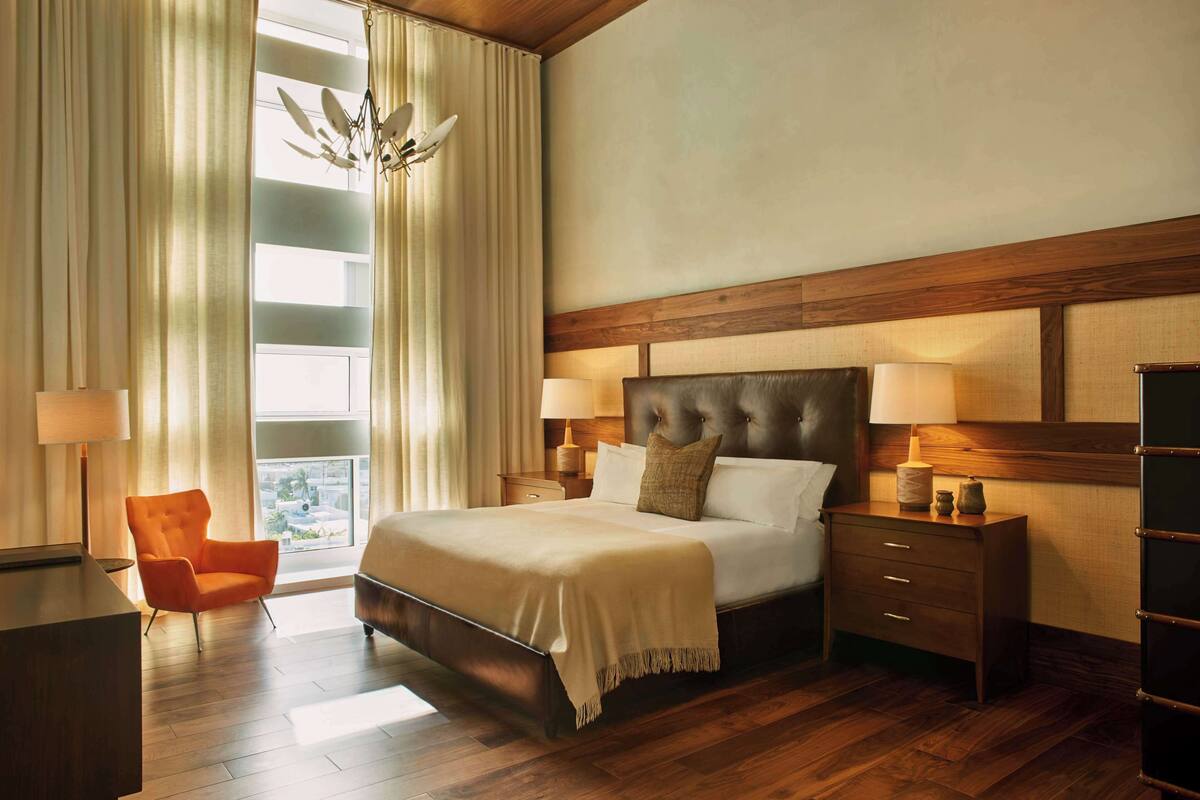 A hotel room with a large bed, wooden headboard, two bedside tables with lamps, a nightstand, a chair, and a view of the outdoors through a large window with curtains.