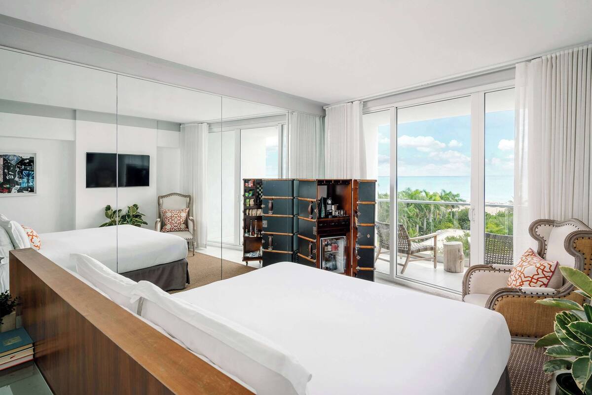 A hotel room with two beds, a large window with a view of the ocean, a bookshelf, and a seating area with a chair and a table.