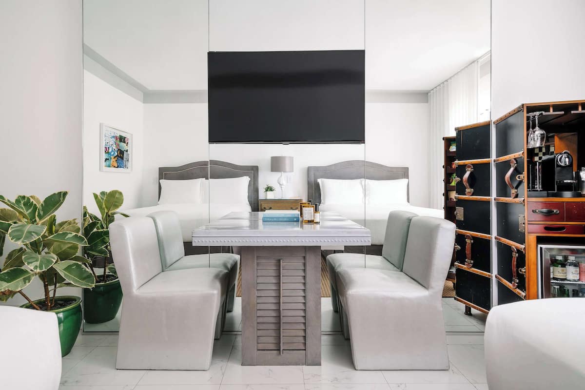 A modern hotel room with a large flat-screen TV mounted on the wall, a dining table with two chairs, a bookshelf, and two potted plants.