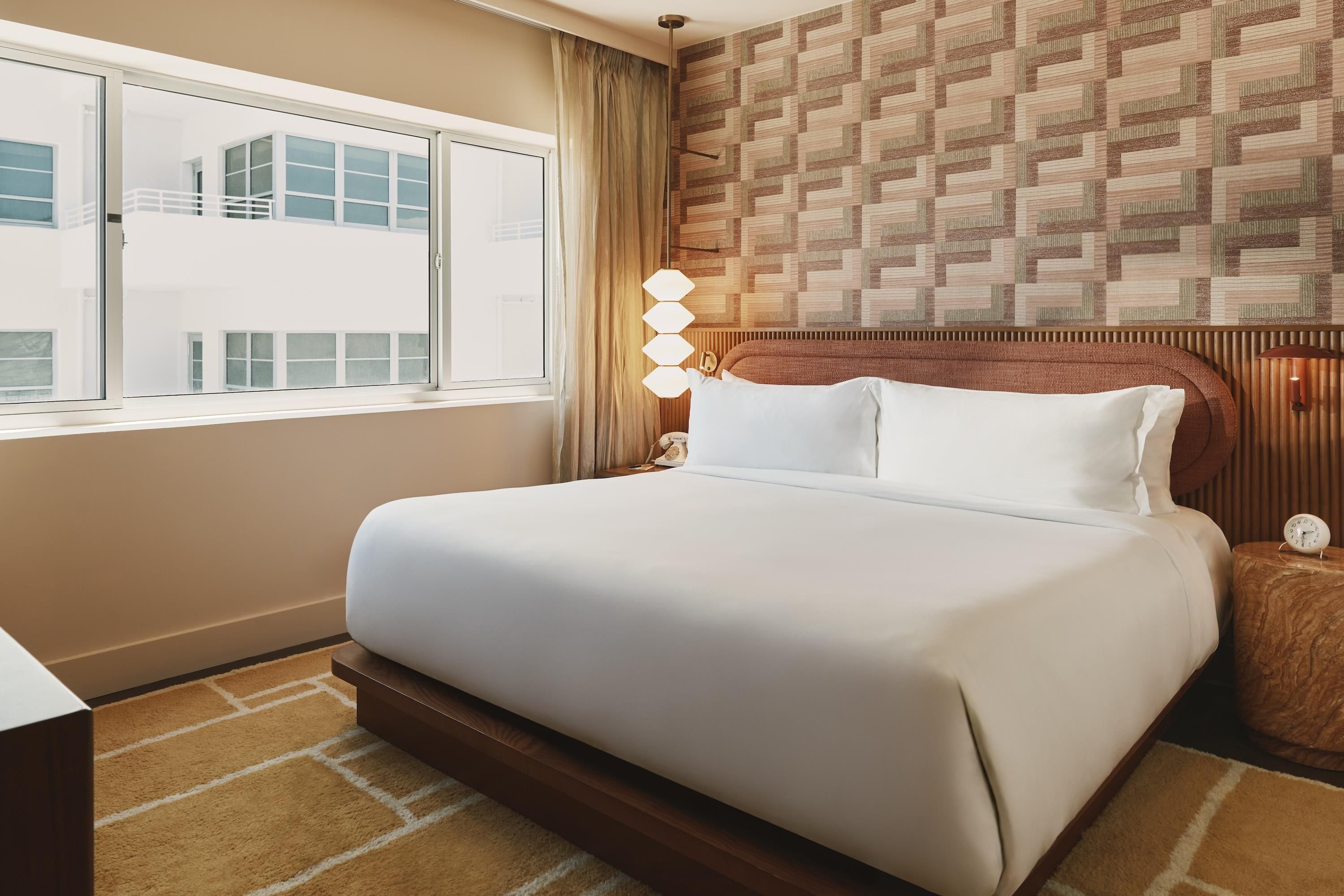 Habitación de hotel con una cama grande, una ventana, un papel tapiz estampado y una mesita de noche con una lámpara.