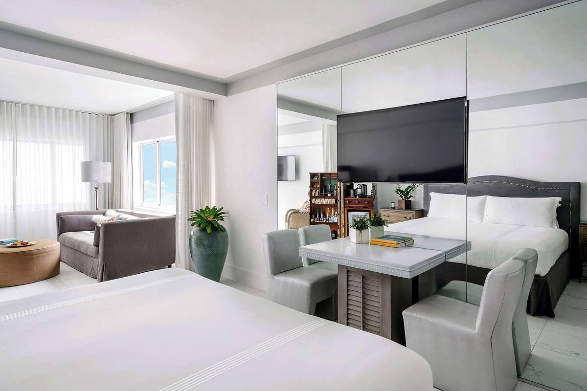 A modern hotel room with a large bed, a desk with a chair, a sofa, a small table, and a large mirror.